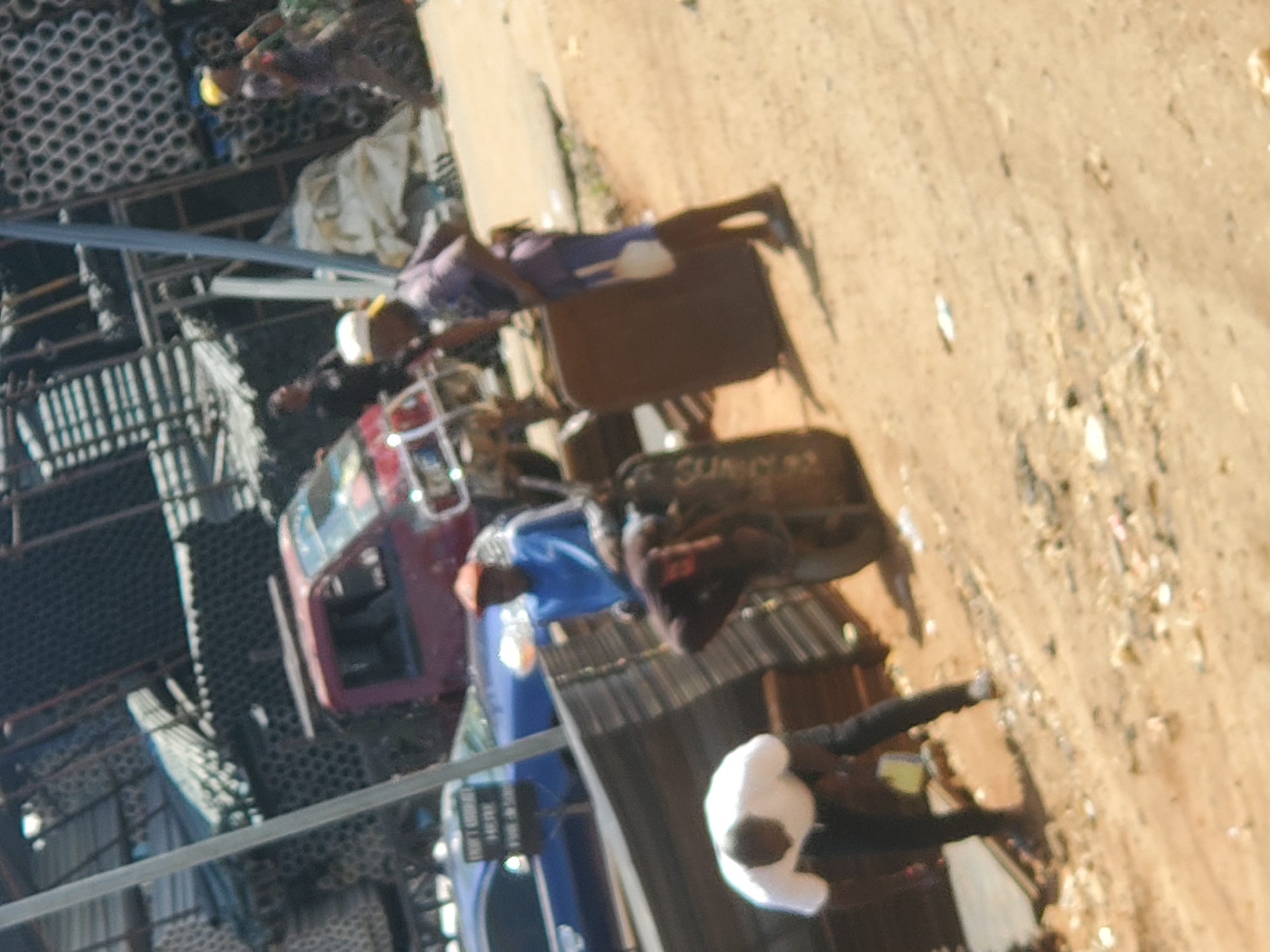 The image depicts an outdoor scene in Fegge, Nigeria. A red vehicle with a metal cage structure is visible. A blue truck is also present with what appears to be signage in English. People are visible in various postures, including standing and moving. One person is wearing a white head covering. Building materials, possibly including pipes, are stacked in the background. The ground is dusty or sandy.