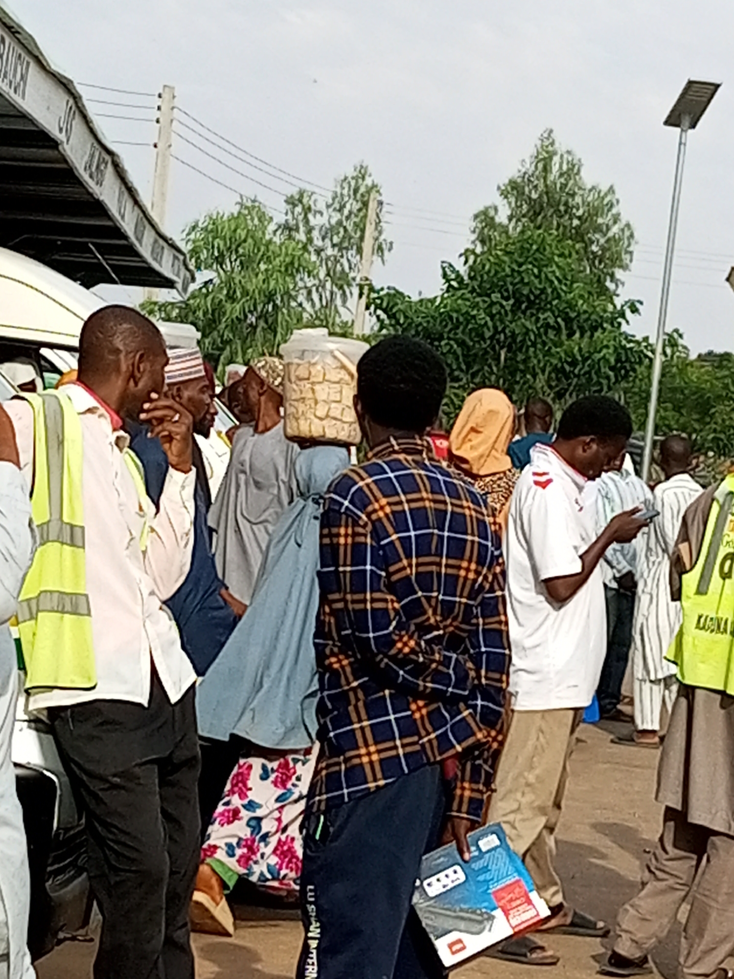 This media file depicts a group of people gathered outdoors, likely in a public space in Gombe, Nigeria. The scene appears to be a busy area, possibly a market or transportation hub, indicated by the presence of a vehicle on the left and a covered structure with signage overhead. The sky is overcast, suggesting a cloudy day.
Several individuals are visible, with a man in the foreground wearing a high-visibility yellow and green vest over a white shirt. He is looking towards another man in a blue shirt and a white and red striped cap, who is gesturing with his hand. Further back, other people are seen in various traditional and modern attire, including flowing robes and head coverings. One person is carrying a large, transparent container filled with what appears to be food items, possibly snacks or pastries. Another individual is focused on their mobile phone.
The overall atmosphere seems to be one of a casual gathering or wait, with people engaged in conversation or simply standing around. The focus is on the human subjects and their interactions within this setting. The presence of electrical poles and wires suggests an urban or semi-urban environment. The clothing styles and the general ambiance point towards a daytime setting. The person in the foreground holding a package with visible text suggests a commercial or transactional activity might be occurring. The visible text on the package is partially obscured but includes what appears to be brand names or product information.