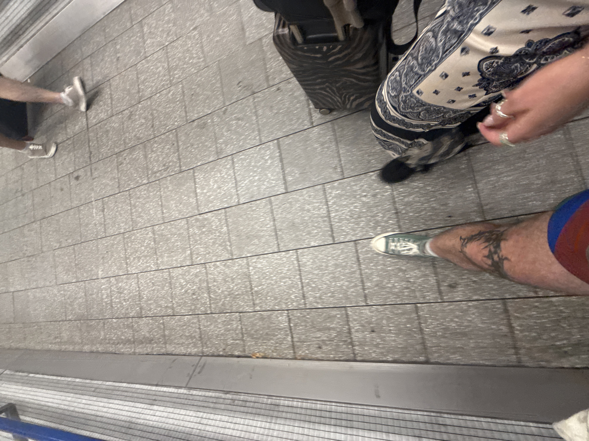 The image depicts a scene in an urban area, likely a transportation hub such as a subway station. The ground is paved with rectangular gray tiles. A partially visible blue railing borders the bottom of the image. Several individuals are present in the photo. One person is wearing a long, patterned dress. Another person is wearing a red and blue swim suit and green sneakers with a tattoo visible on their lower leg. A piece of luggage is also visible. The scene appears to be a waiting area, given the presence of luggage and standing individuals.