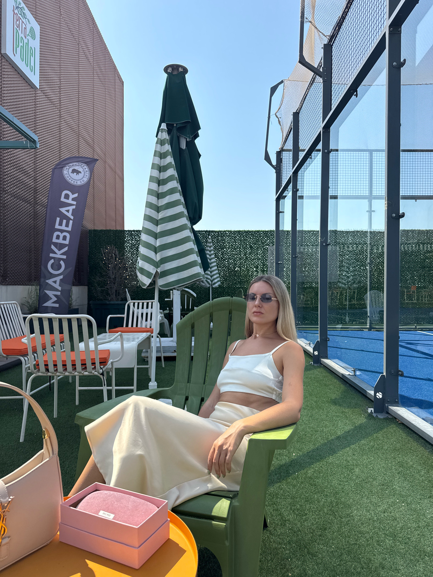 A blonde woman with sunglasses sits in a green chair on a sunny day. She is wearing a white crop top and a cream-colored skirt. In front of her is a small orange table with a pink gift box and a beige handbag. Behind her is a padel court with a blue surface and glass walls. There are also umbrellas and chairs in the background. The location is Terra Padel in Antalya, Turkey.