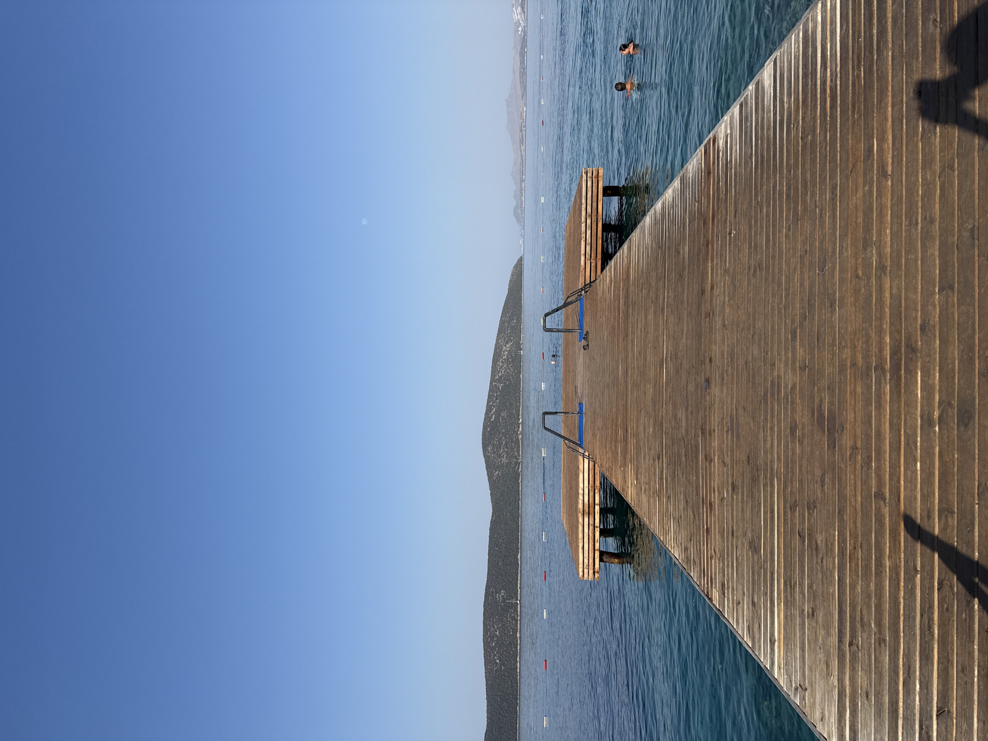 This is a vertical, eye-level, outdoor, full shot photograph taken on a sunny day in Bodrum, Turkey, featuring a wooden pier extending into calm, clear blue water. The pier takes up the majority of the foreground and middle ground. It is constructed of weathered wooden planks running horizontally. At the end of the pier, there are two sets of metal handrails with blue steps leading into the water. Two people are visible swimming in the sea to the right of the pier. In the distance, rolling hills covered in green foliage form the horizon. The sky above is a clear, vibrant blue, with a faint crescent moon visible in the upper right quadrant. The water is a serene blue-green, with ripples indicating gentle movement. The scene evokes a sense of tranquility and summer leisure.