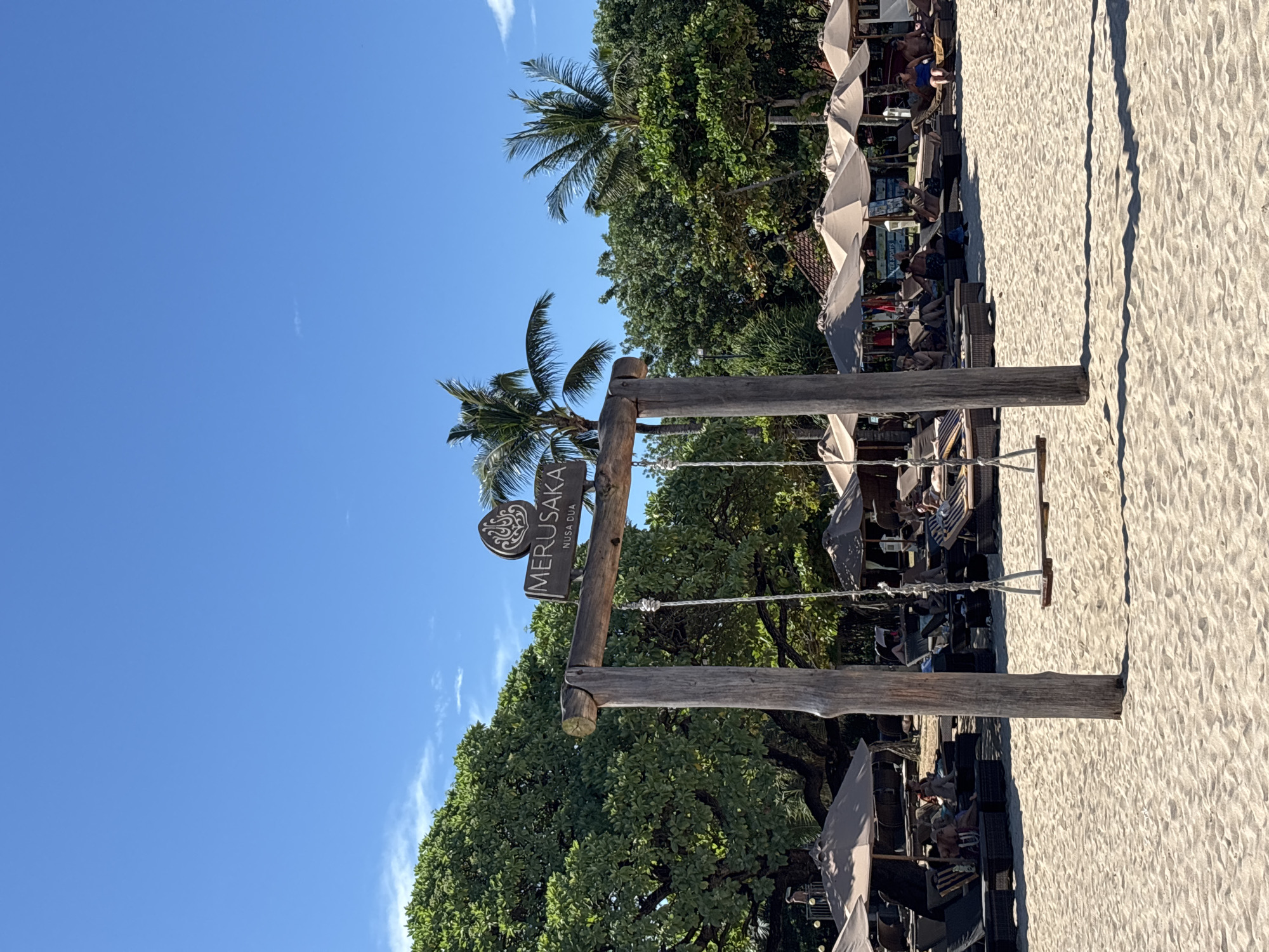 The image captures a beach scene in Nusa Dua, Indonesia. A signpost indicates the location as "Merusaka Nusa Dua." The scene includes a wooden swing set and outdoor seating areas with tables and umbrellas. People are seated and lounging in the seating areas. The background features lush green trees and a clear blue sky. The foreground is a sandy beach.