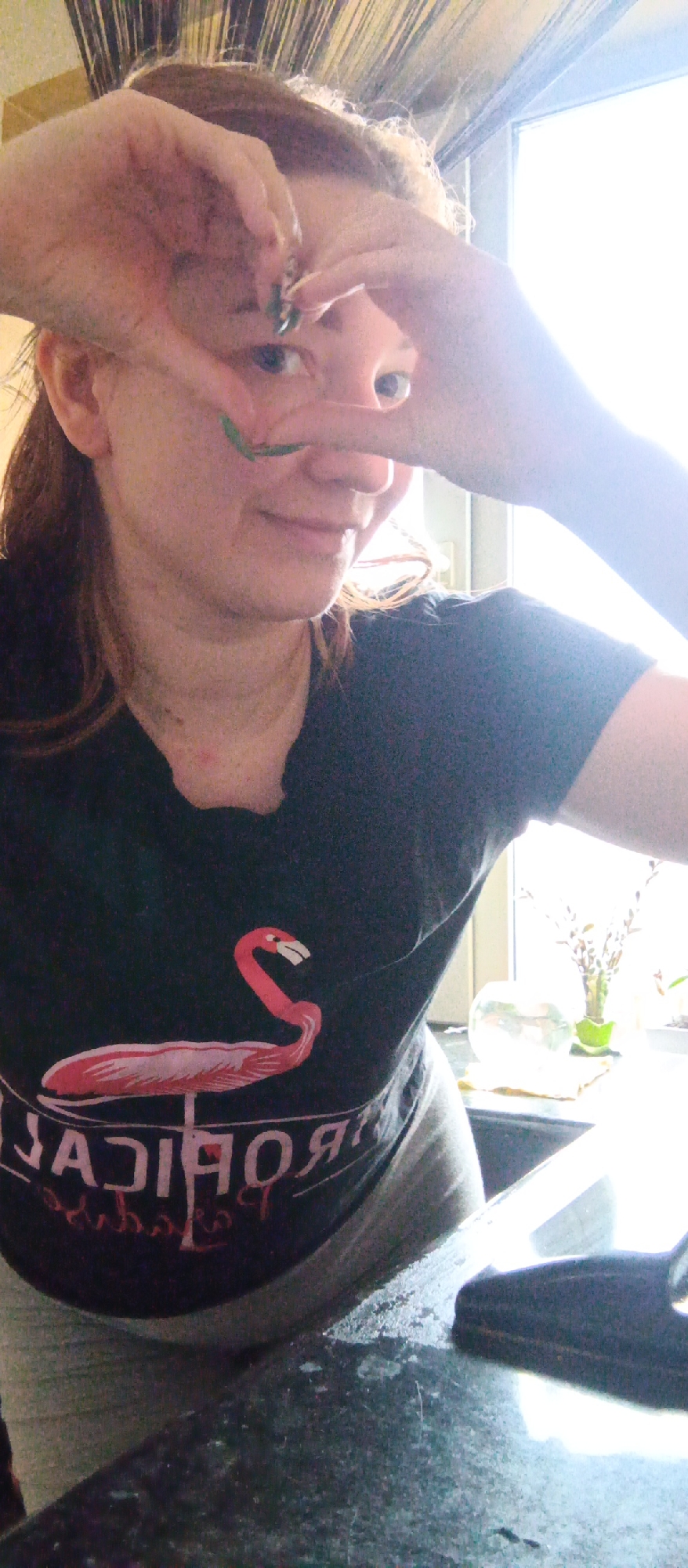 A person is indoors, posing for a selfie. They are wearing a black t-shirt with a pink flamingo design. The person has fair skin and is holding their hands up near their face, with one finger touching their nose. Green markings are visible on their face and nails. Behind the person is a window with a bright, diffused light source, and a glimpse of plants or greenery is visible on the windowsill. The countertop in front of the person appears to be dark-colored. The location is unknown.