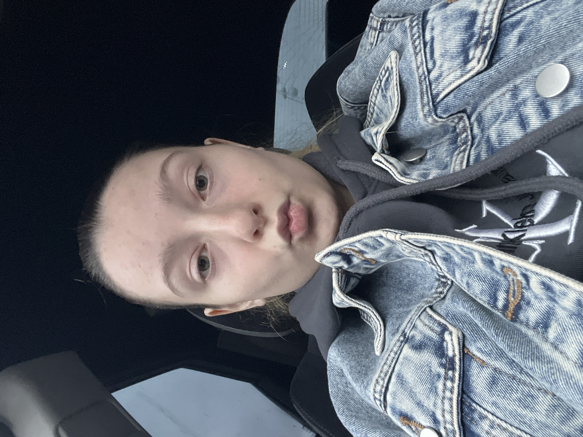 A young individual with fair skin and light brown hair pulled back is posing for a selfie in what appears to be a car. The person is wearing a grey hooded sweatshirt under a light blue denim jacket. The interior of the car is mostly black, with a window visible in the background. The location is not identifiable as Varna, Bulgaria, from the visual information provided. There is nothing distinctive or recognizable about the background or objects to associate the scene with the city.
