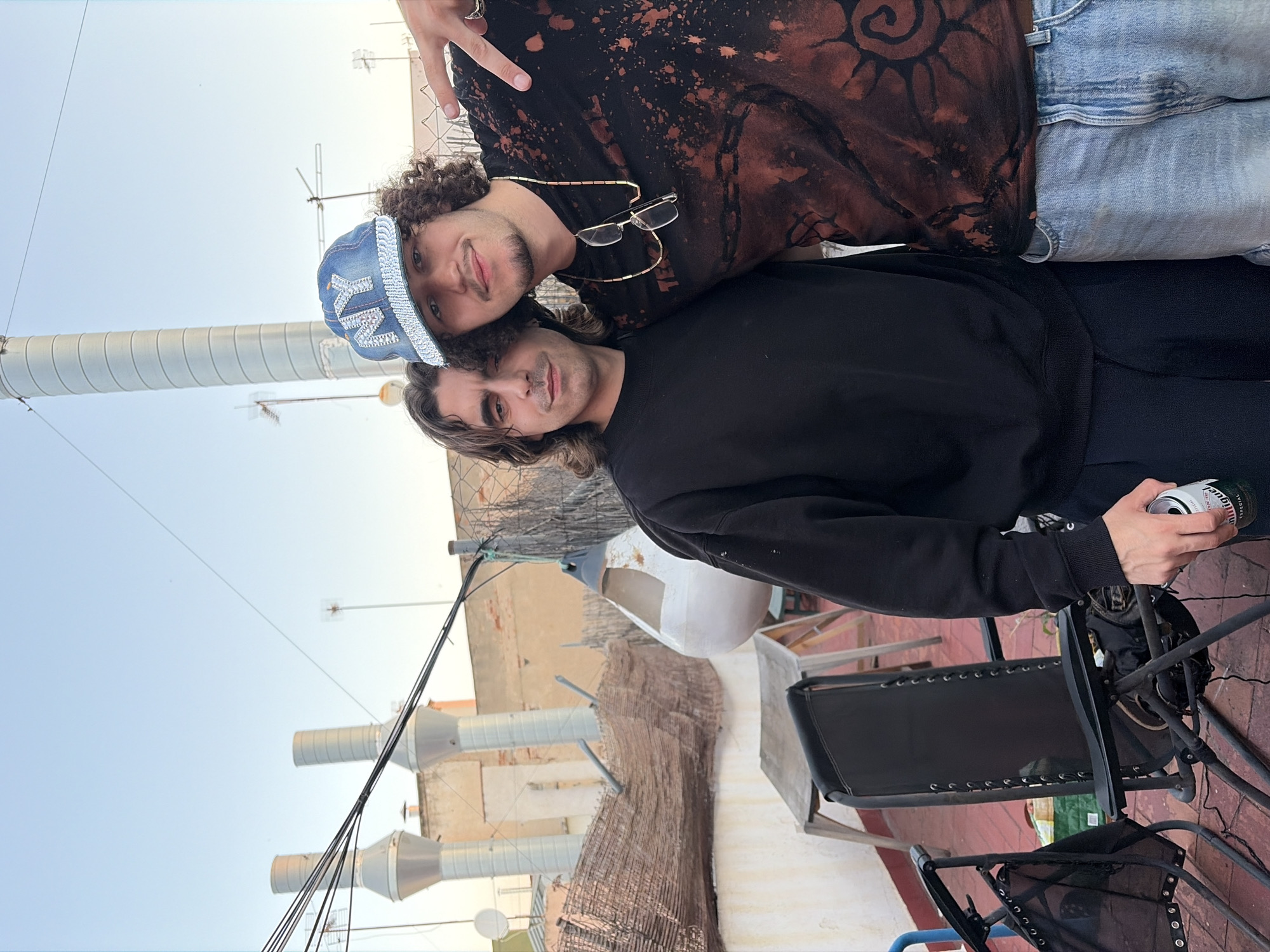 Two men are posing together on a rooftop. One man wears a black shirt with a brown circular design, blue jeans, and a blue baseball cap with "NY" on it, also having a pair of glasses hanging on his shirt. The other man wears a black sweatshirt. The background shows urban elements, including chimneys, satellite dishes, and buildings, indicating the location is likely the city of Barcelona, Spain. One of the men is holding a drink. The environment is a rooftop setting with outdoor furniture.
