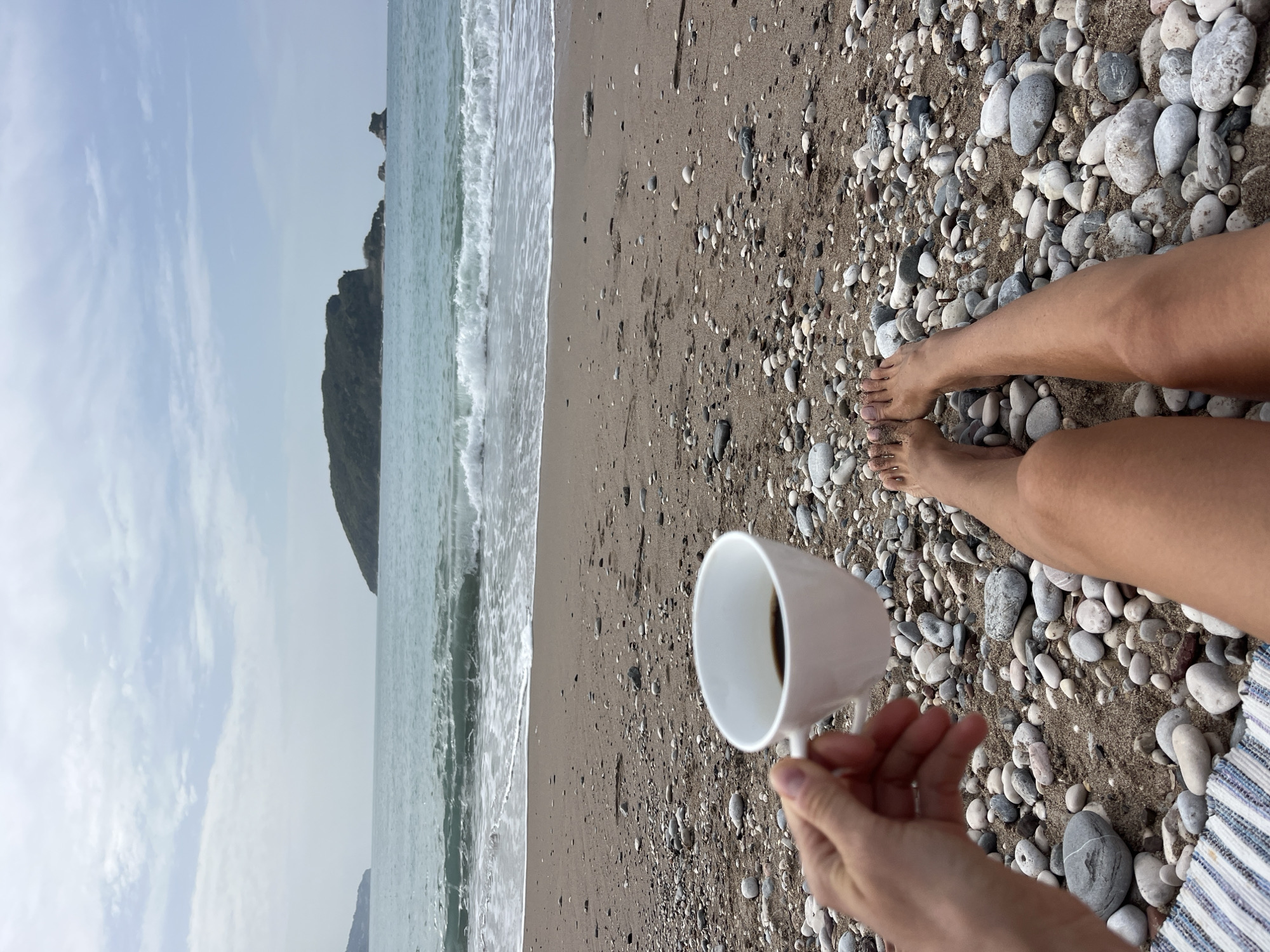 A medium shot captures a beach scene. The sky is overcast, and the calm sea features gentle waves meeting a sandy beach. A rocky hill is visible in the distance.
In the foreground, a person sits on the beach, holding a white cup. The beach consists of sand and small pebbles. The person's bare legs and feet are visible, resting on the pebbles. A portion of striped fabric rests beside the right leg.
The image likely depicts the beach, named Mogren beach, in the city of Budva, Montenegro.