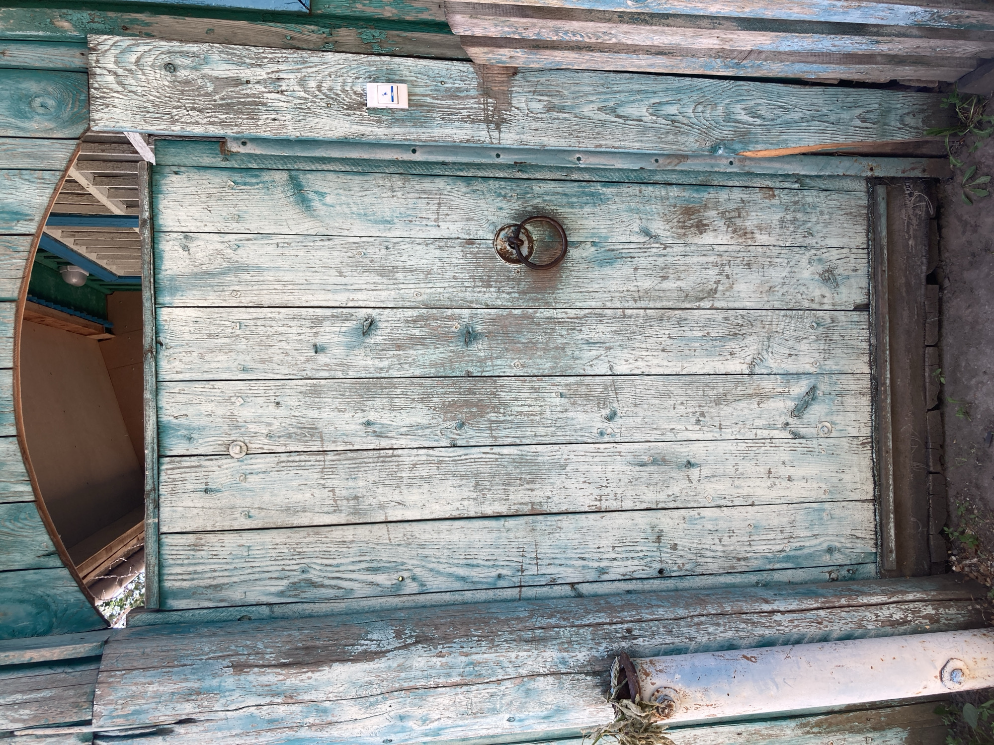 The image shows a weathered, pale blue wooden door with a circular metal door knocker. The door is framed by a similarly colored wooden structure that appears to be part of an entryway or gate. Above the door, there is an arched opening revealing a glimpse of a wooden ceiling and some greenery outside. To the left of the door, a rusted metal pipe stands upright, with dead foliage protruding from its top. The ground in front of the door is covered with dirt and some sparse vegetation. The overall impression is one of age and disuse. The location is described as Rakity, Russia.