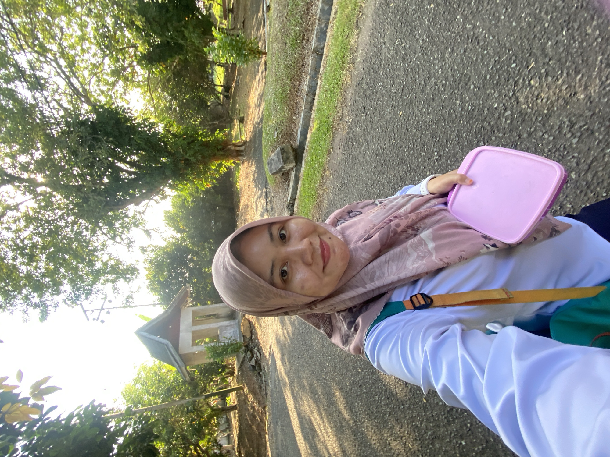 A young woman wearing a pink hijab and a white long-sleeved shirt takes a selfie outdoors in Banjarbaru, Indonesia. She holds a pink lunchbox in her right hand. A green backpack with an orange strap is visible over her left shoulder. She is standing on a paved road with sunlight casting shadows. In the background, there is a small, open-sided pavilion with a green roof, surrounded by trees and lush greenery. The scene appears to be a park or a campus area.