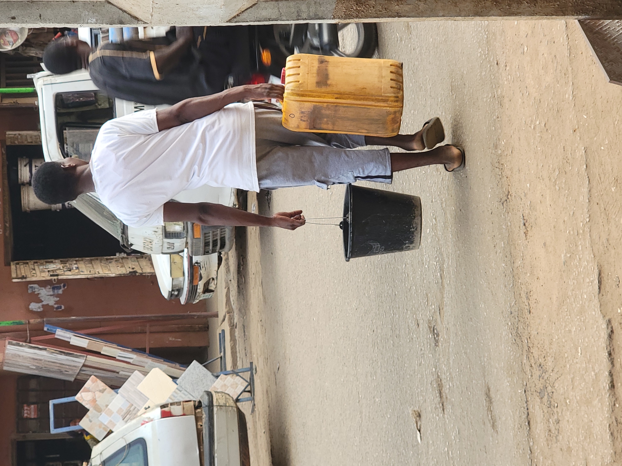 The image captures a man in Maiburiji, Nigeria, walking on a street with a yellow jerrycan in one hand and a black bucket in the other. He is wearing a white t-shirt, grey shorts, and sandals. He is walking on the right side of the street on a light brown surface. A white vehicle with tiles on top is visible behind him. Another man wearing a black shirt can also be seen next to the first vehicle. The environment appears to be an urban street.