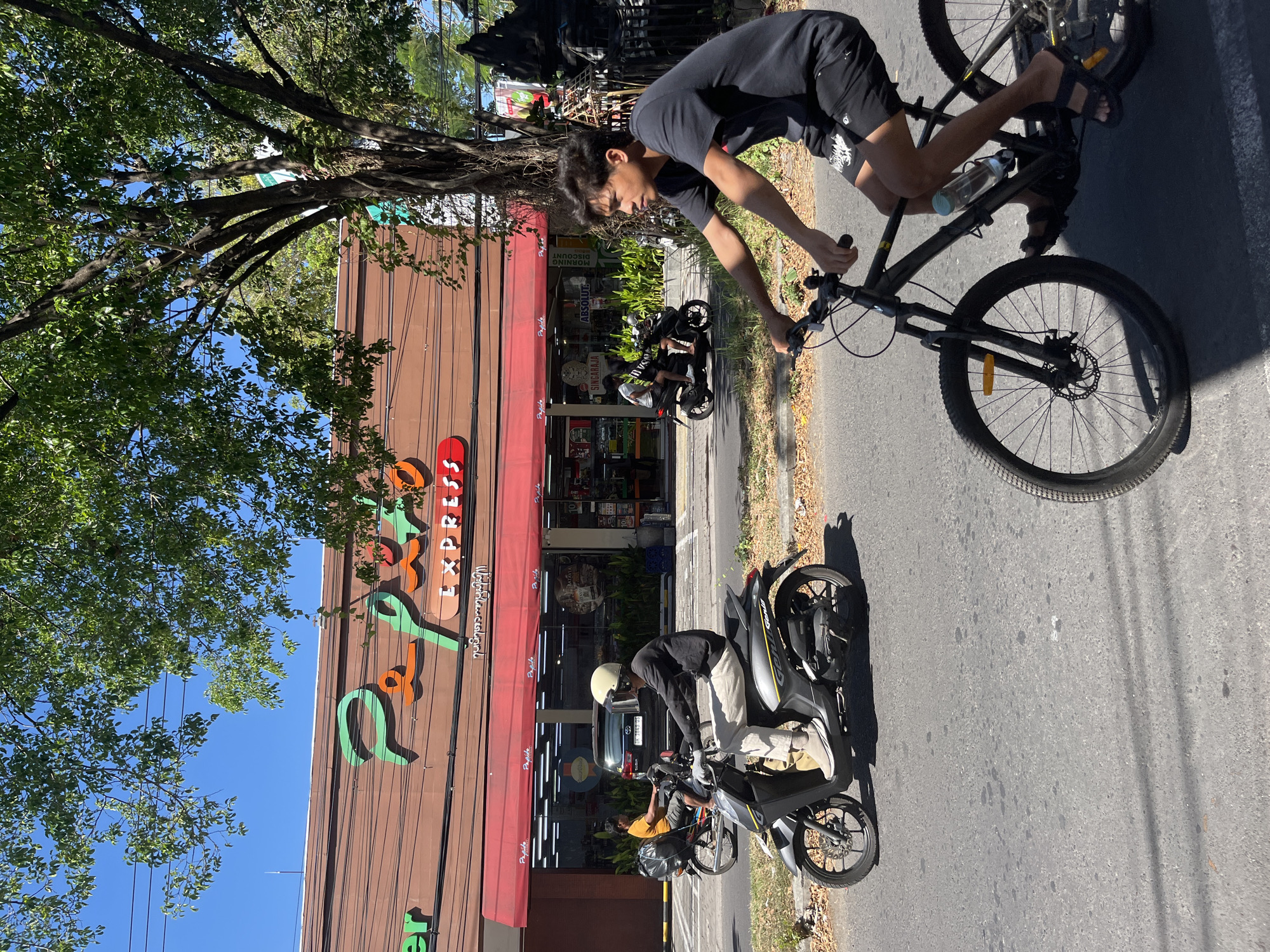 The image depicts a street scene in Kuta Selatan, Indonesia, on a sunny day. In the foreground, a young man with dark hair, wearing a black t-shirt and black shorts, is riding a black bicycle. He is wearing dark sandals and has a water bottle attached to his bicycle frame. His bicycle has a front disc brake and a water bottle cage. To his left, a gray scooter is being ridden by a person wearing a white helmet with a dark visor, a dark long-sleeved shirt, and light-colored pants. The scooter has a black rear storage box. Further behind them, another scooter is visible with two people on it.
In the background, a building with a brown facade displays a prominent green and orange logo that reads "Papua Express". Red awnings cover the storefront windows, through which various products and displays are visible, including signage that says "Batavia" and "Singaraja". Several trees with lush green foliage line the street, casting shadows on the asphalt road. Electrical wires are strung overhead. The sky is clear and blue. The overall impression is a typical street scene in a busy, tropical urban environment.