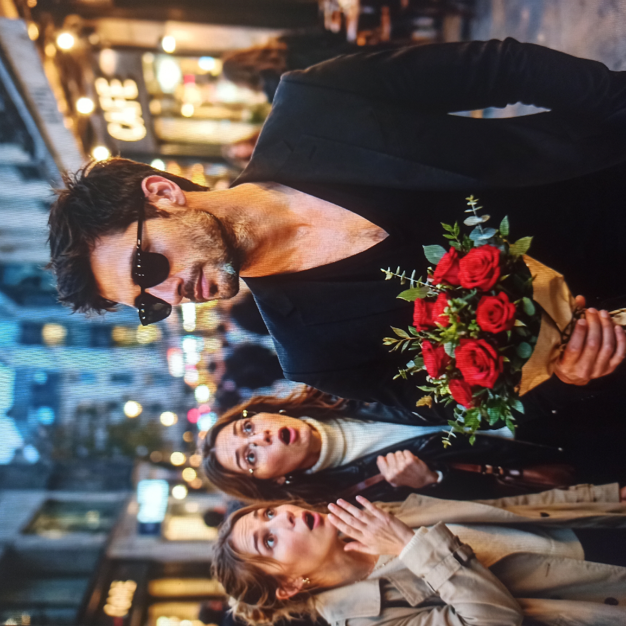 The image shows a man holding a bouquet of red roses in front of two women. The man is wearing sunglasses and a black jacket over a black v-neck shirt. The women are looking at something to their right with surprised expressions. The woman on the left is wearing a beige trench coat and the woman on the right is wearing a black leather jacket and a white turtleneck. They appear to be on a city street at dusk, with blurred lights and buildings in the background. One building has a sign that reads "CAFE". The image does not depict any recognizable landmarks or features specific to Nizhny Novgorod, Russia.