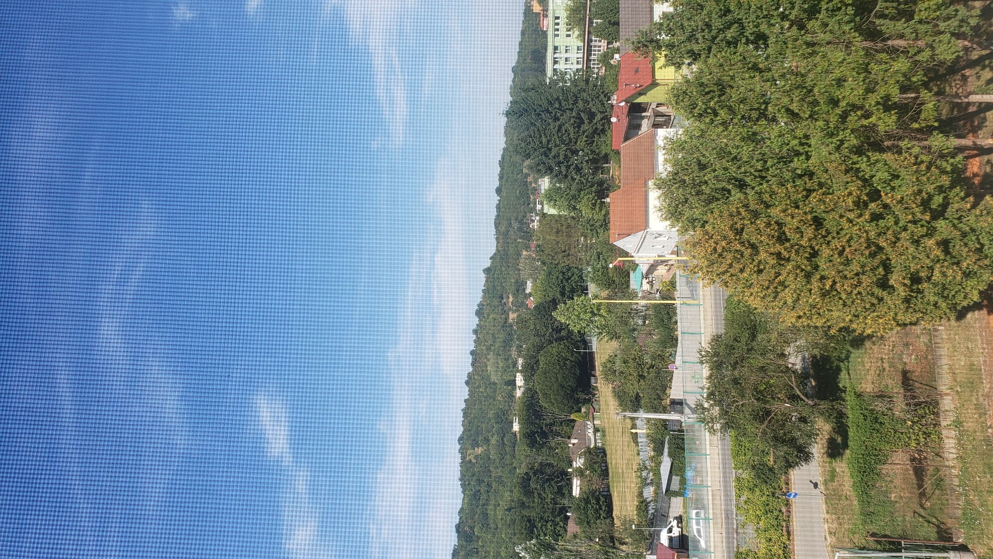 The image depicts an aerial view of a residential area in Prešov, Slovakia, on a clear day. The sky is a vibrant blue with scattered white clouds, and a noticeable grid pattern from a window screen overlays the entire image.
In the foreground, several large trees with green and yellowing foliage dominate the view. Beyond the trees, a road is visible, with a white van parked on the left side of the frame. A fence runs along the edge of the road, separating it from a grassy area and several houses with red and white roofs. In the mid-ground, more houses and trees are scattered across the landscape, with a larger green building visible in the distance on the right.
The background is characterized by a densely wooded hill that rises behind the residential area, suggesting the city is situated near a natural landscape. The overall impression is one of a peaceful, suburban environment bathed in sunlight.