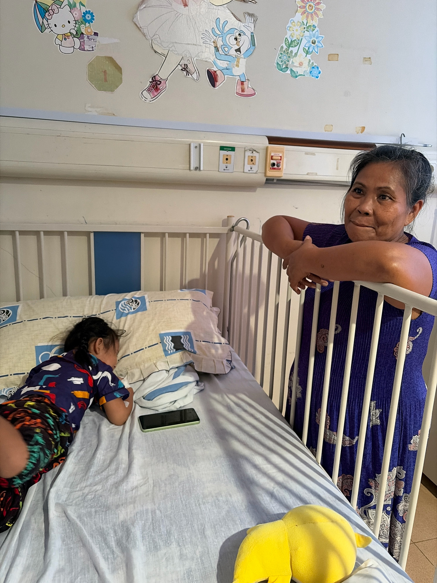 In a room decorated with cartoon stickers, a woman stands by a crib, her arms resting on the railing. Inside the crib, a child lies asleep on a pillow, wearing a colorful shirt and shorts. A cell phone is placed on the bed next to the child, and a yellow stuffed toy is visible in the foreground. The room appears to be a hospital room, possibly in Tanjung Pinang, Indonesia, suggested by the style of the crib and medical equipment visible on the wall.