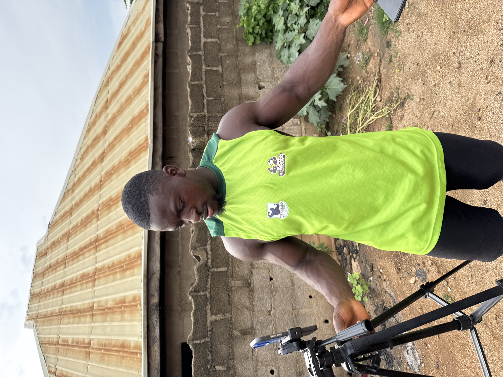 In Akwanga, Nigeria, a Black man with short hair and a beard stands outdoors in front of a brick wall and a rusty metal roof. He is wearing a bright green sleeveless shirt with green trim and black shorts. On his shirt, there are two logos; the left one is a white shield with a blue outline containing a stylized figure and the text "SUPA", and the right one is a similar shield with a blue outline containing two stylized figures and the text "KOMANDO ENERGY DRINK". The man is looking down at a tripod with a phone mounted on it, positioned to his left. His left arm is extended towards the tripod, and his right arm is extended to his right, holding a black smartphone. The ground is covered in dirt and gravel, with some green vegetation visible in the background. The sky is overcast.