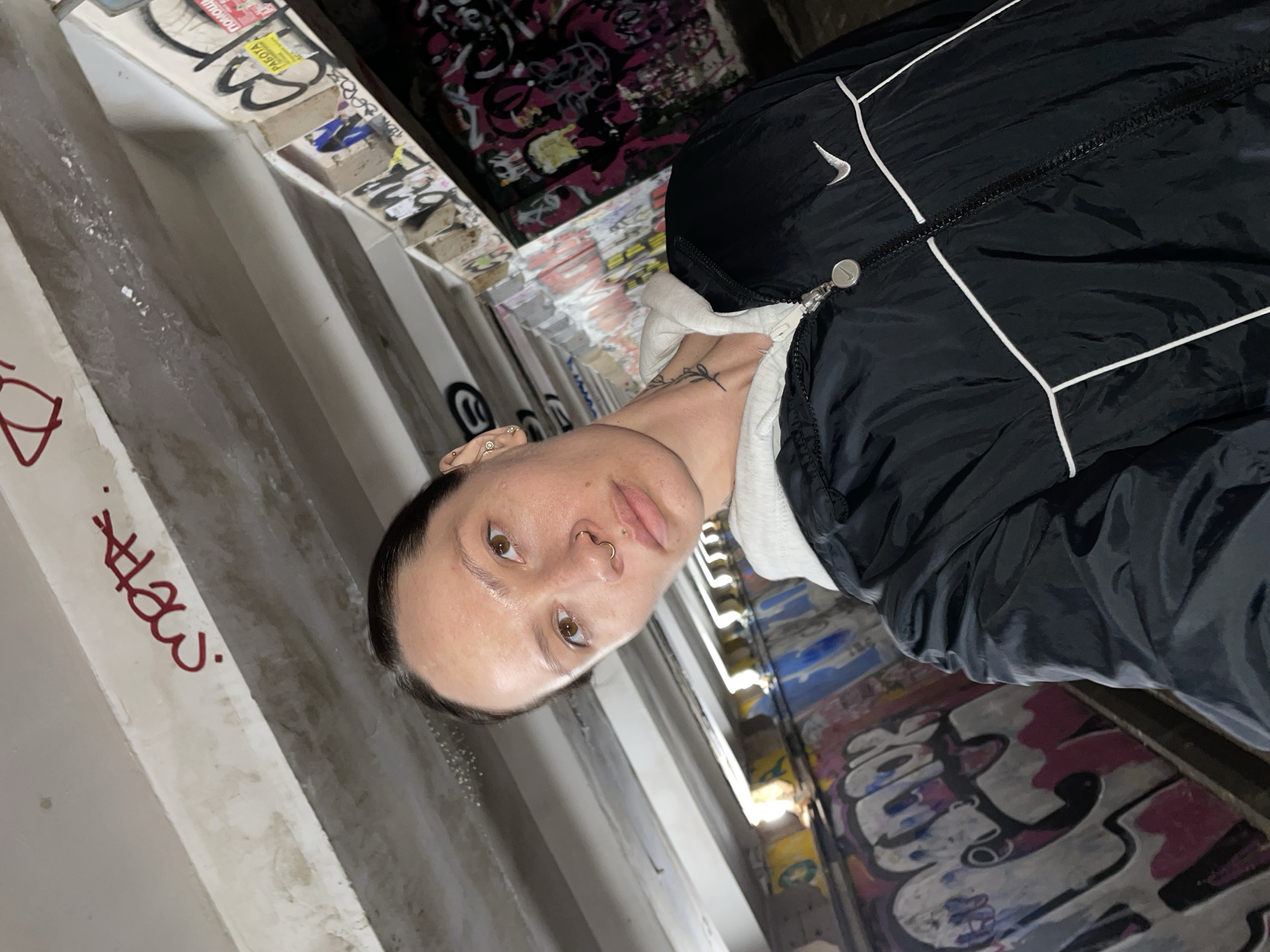A medium, eye-level shot shows a person with short, dark hair, light skin, brown eyes, and a nose ring standing in front of a wall covered in graffiti. The person is wearing a black jacket with white stripes and a light-colored hoodie underneath. The graffiti on the wall is multi-colored, with shades of pink, blue, yellow, and white visible. To the left of the person is a concrete structure with red writing. The location appears to be an urban environment, possibly a tunnel or underpass. The graffiti suggests an area known for street art, although the exact location in Izhevsk, Russia, cannot be determined from this image alone.