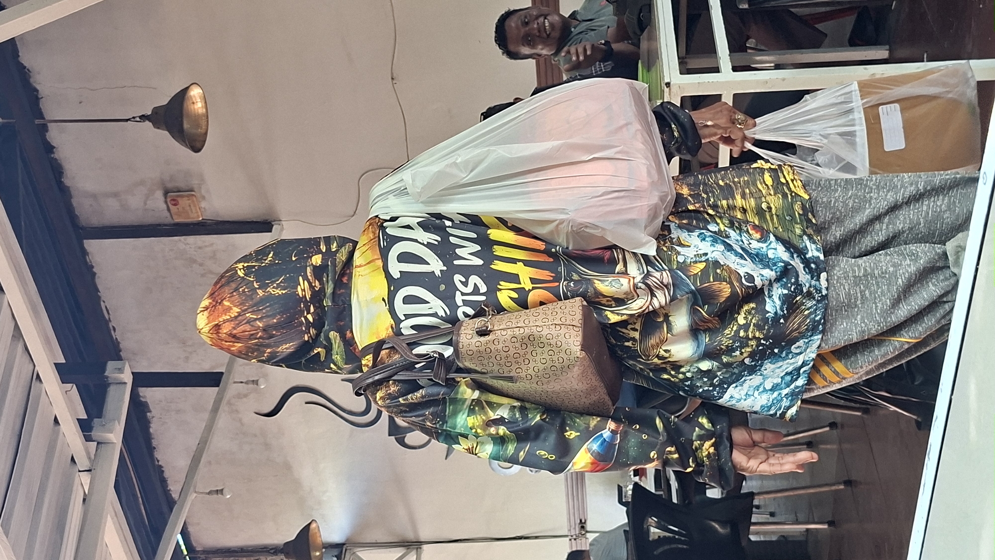 The image depicts the rear view of a person wearing a hooded jacket with a vibrant, multi-colored print featuring fish and text that partially reads "GOOD DAY SPORTS WITH SHI...". The person is carrying a brown monogrammed handbag on their left shoulder and two plastic bags. One white plastic bag, filled with orange items, is slung over the right shoulder, and a second clear plastic bag containing a brown granular substance is held in the right hand. The person is standing indoors, with elements of what appears to be a restaurant or food court visible. In the background, a man is seated at a table, smiling and giving a thumbs-up gesture. The setting includes tables, chairs, ceiling lights, and a textured wall. The specific location within Lhokseumawe, Indonesia, is not identifiable from the visual information provided in the image.