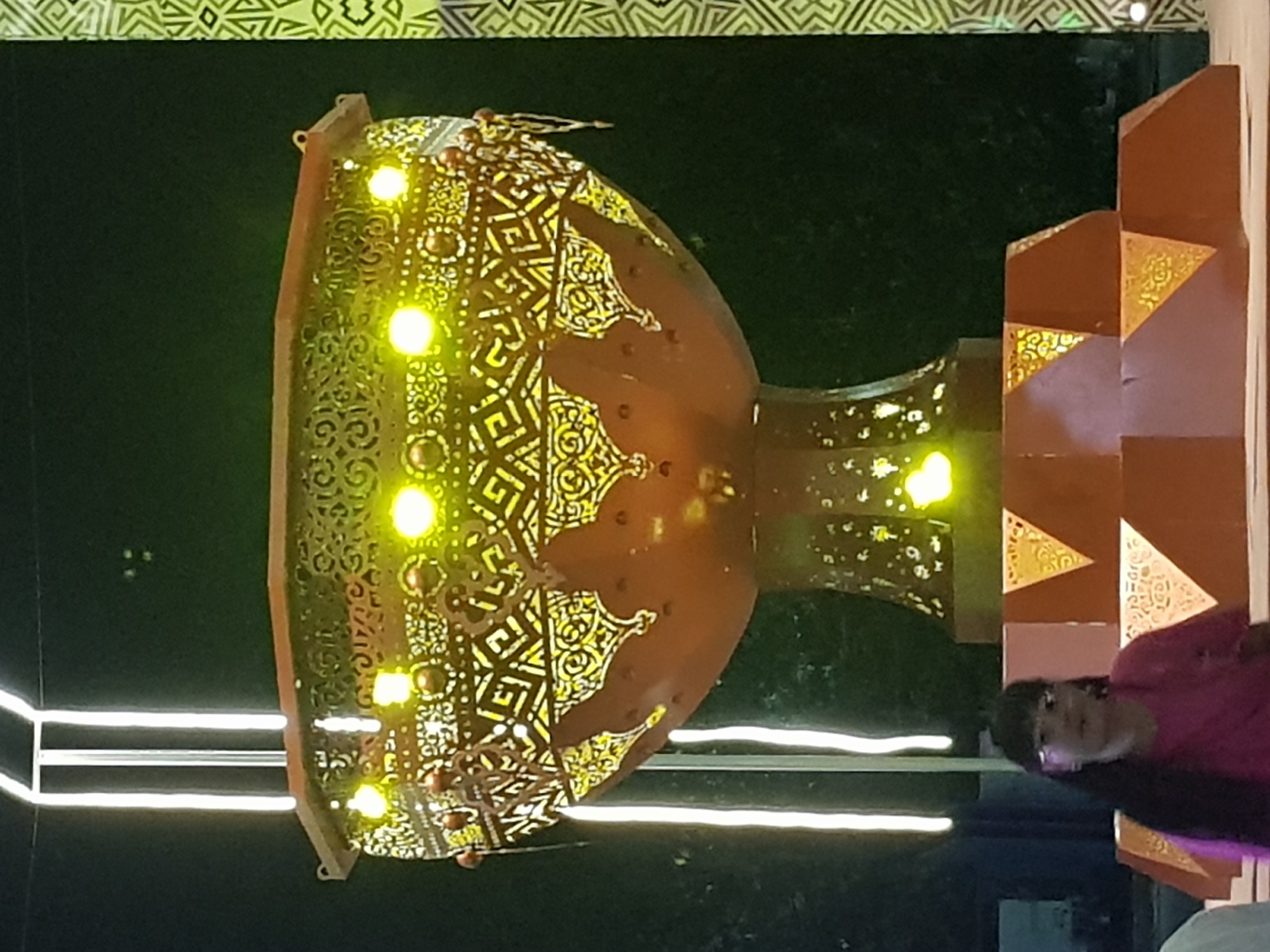 A young girl with dark hair, wearing glasses and a pink t-shirt, stands in front of a large, ornately decorated, copper-colored bowl illuminated by yellow lights. The bowl is supported by a pedestal decorated with geometric patterns. The scene appears to be outdoors at night, with vertical white lights visible in the background and a dark, textured wall on the right side of the frame. The overall setting suggests a festive or decorative display, possibly in the city of Kurchatov, Kazakhstan.