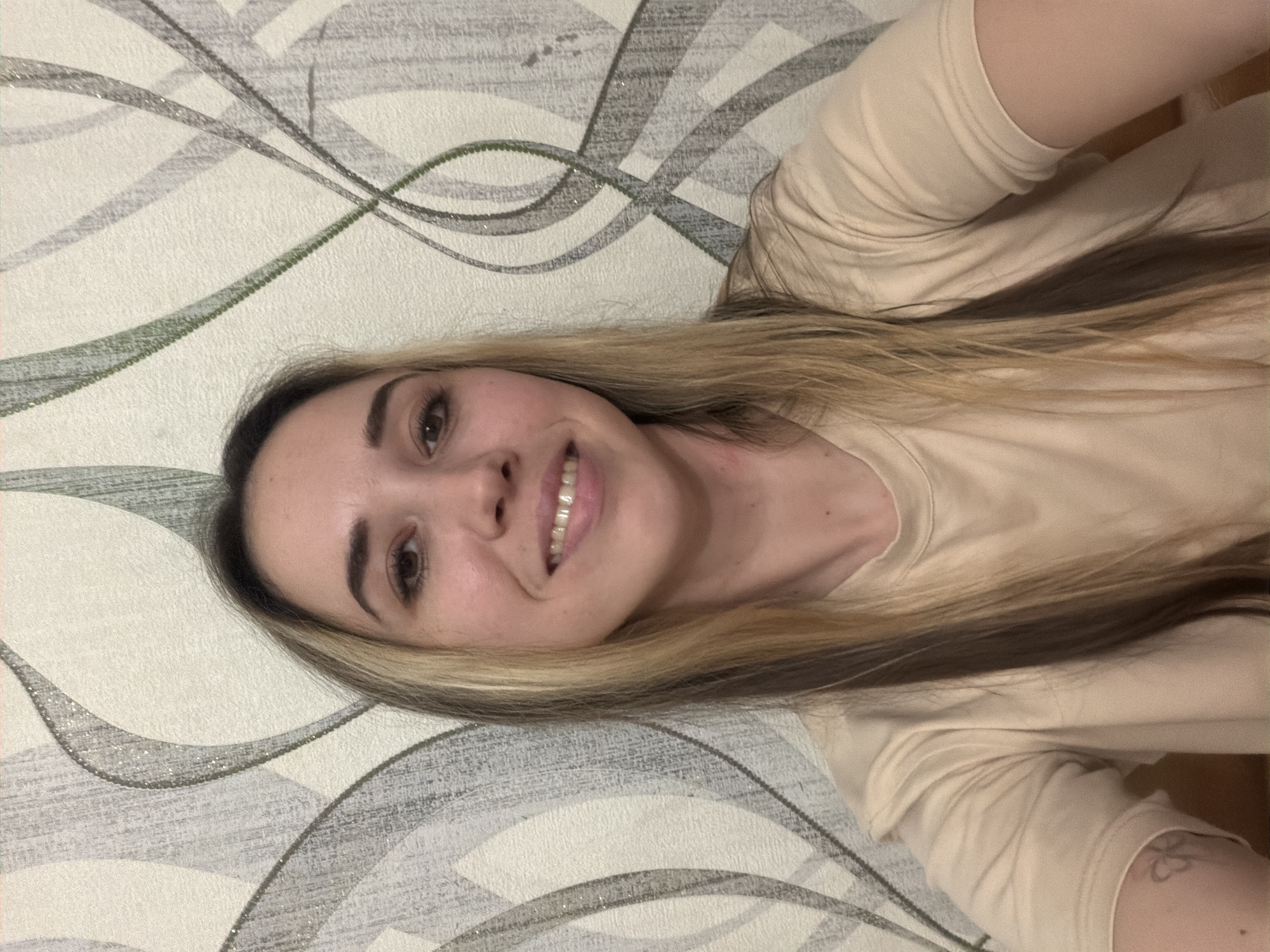 A person is lying down against a wall with a patterned covering. The person has long blonde hair, brown eyes, and is smiling. The person is wearing a light-colored t-shirt. A tattoo is visible on the person's arm. The location is Komsomolskiy, Russia, but there is no evidence of it present in the image.