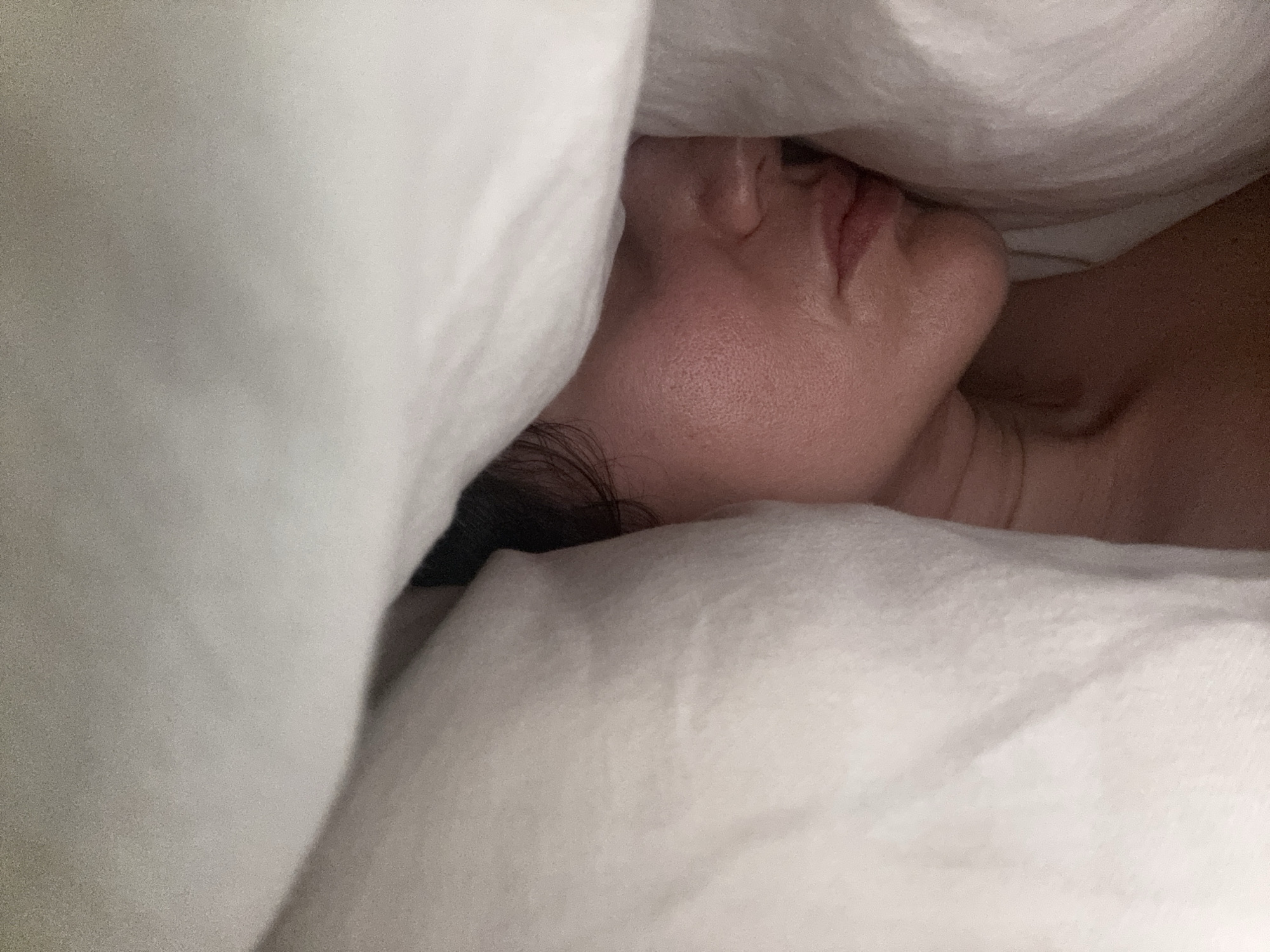 The image captures a person sleeping or resting peacefully, nestled amongst white bedding, likely in an indoor setting in Dubai, United Arab Emirates. The subject's face is shown in profile, with their forehead, closed eye area, nose, and mouth softly illuminated. Dark hair is visible near the back of their head and neck.
The dominant objects in the frame are the soft, white sheets, blankets, or pillows that cocoon the person, creating a sense of comfort and tranquility. The folds and textures of the fabric are visible, adding to the intimate feel of the scene. The lighting is gentle and muted, characteristic of a bedroom environment, suggesting either night or early morning. The overall atmosphere conveys a feeling of deep relaxation and serenity. No text is visible in the image.