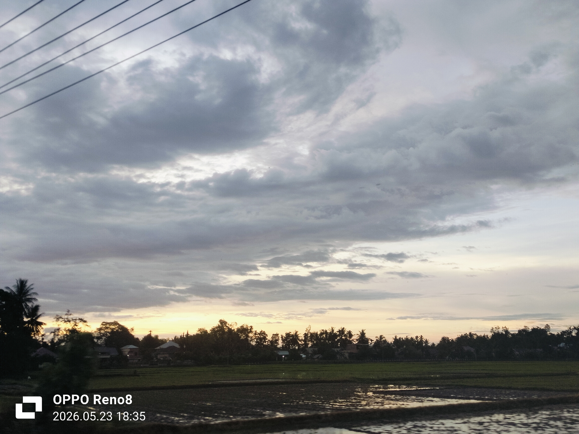 The image depicts a landscape in Kampungjumpa, Indonesia, at sunset. The sky is overcast with grey clouds, revealing a band of golden light along the horizon. In the foreground, a flooded rice field reflects the ambient light. Beyond the field, a dense row of trees and vegetation obscures the houses and structures. Power lines are visible in the upper left corner of the image. The photograph was taken on an OPPO Reno8, as indicated by the watermark, with the date and time stamp showing May 23, 2026, at 18:35. There are no people visible in the image.