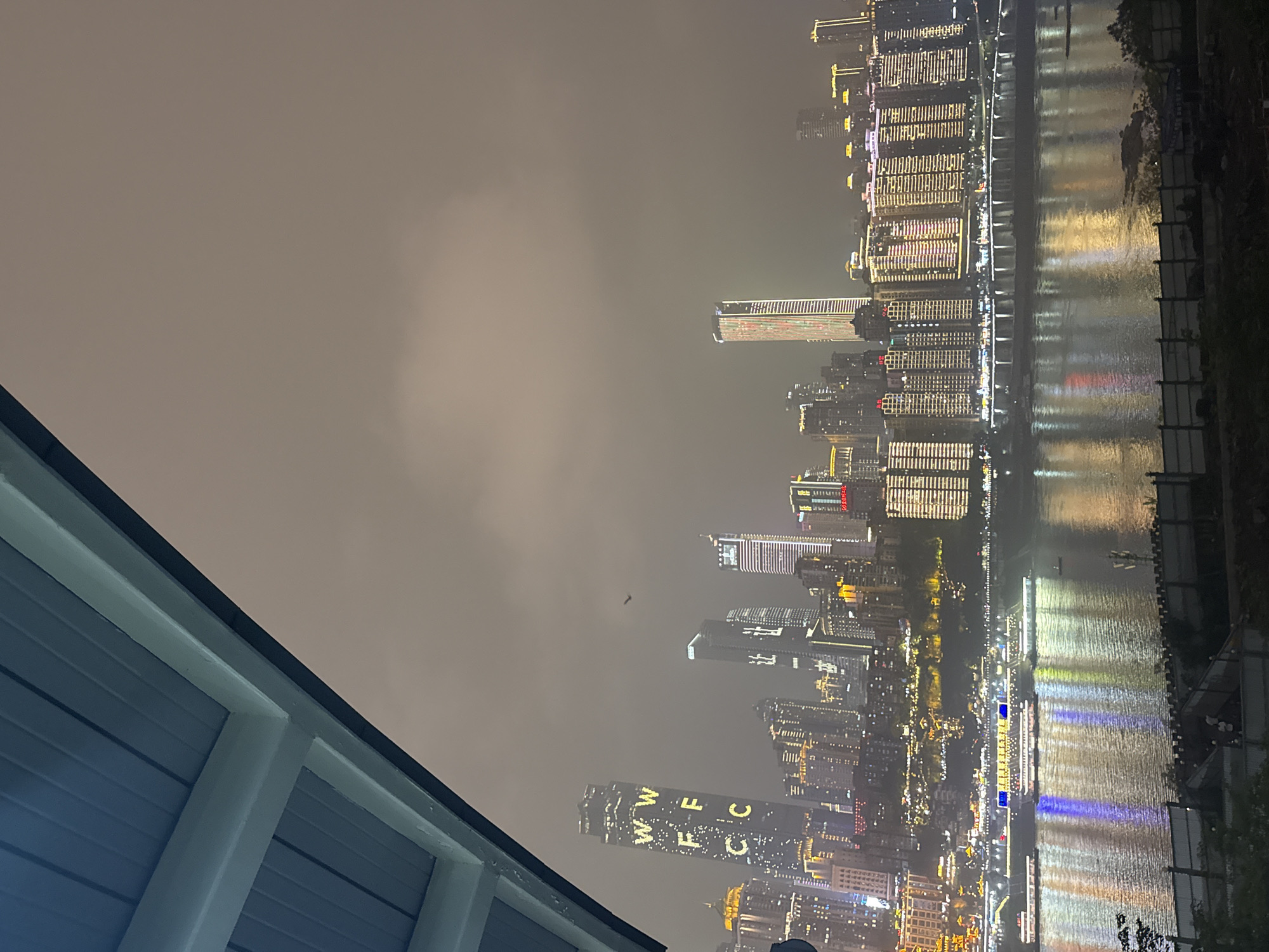The image captures a nighttime view of the city skyline of Yuzhong District, China, featuring numerous illuminated buildings alongside a body of water, possibly the Yangtze or Jialing River. The lights of the buildings reflect in the water, creating a mirrored effect. The sky is overcast, with a hazy or foggy quality muting the brightness of the lights. In the lower left corner is a portion of a building structure, with light blue paneling and white framework. The city's architecture is modern, with many high-rise buildings displaying elaborate lighting designs. One building prominently displays the letters "WWFCC" with lights.