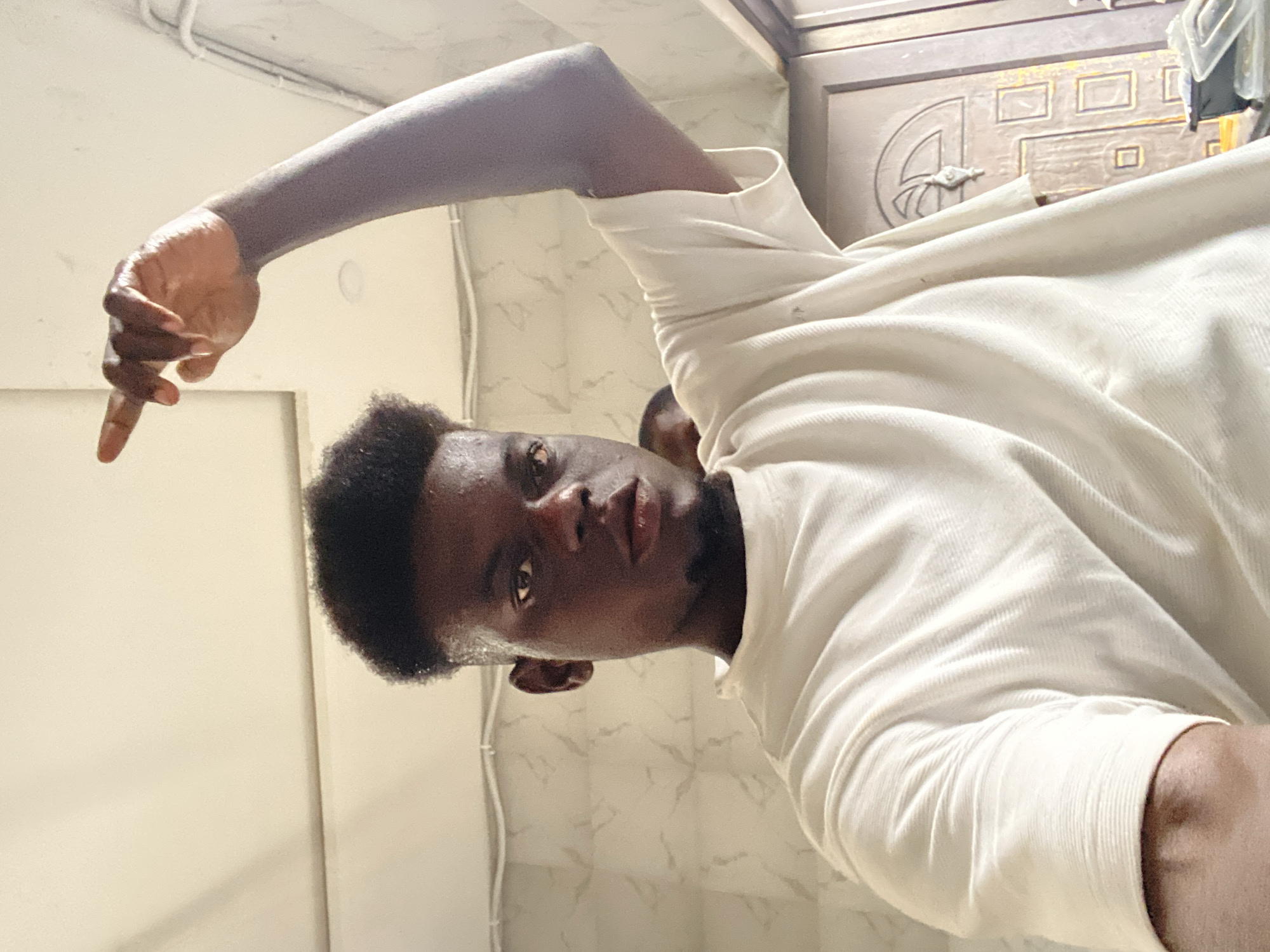 A medium-shot photo from an unusual angle depicts a person with dark skin and short, dark hair. The individual is wearing a light-colored, long-sleeved shirt. Their arm is raised, with their index finger pointing downwards. The background includes a white wall with visible pipes, and a partially visible door with decorative panels. The location appears to be indoors, possibly in the city of Unknown, Unknown.