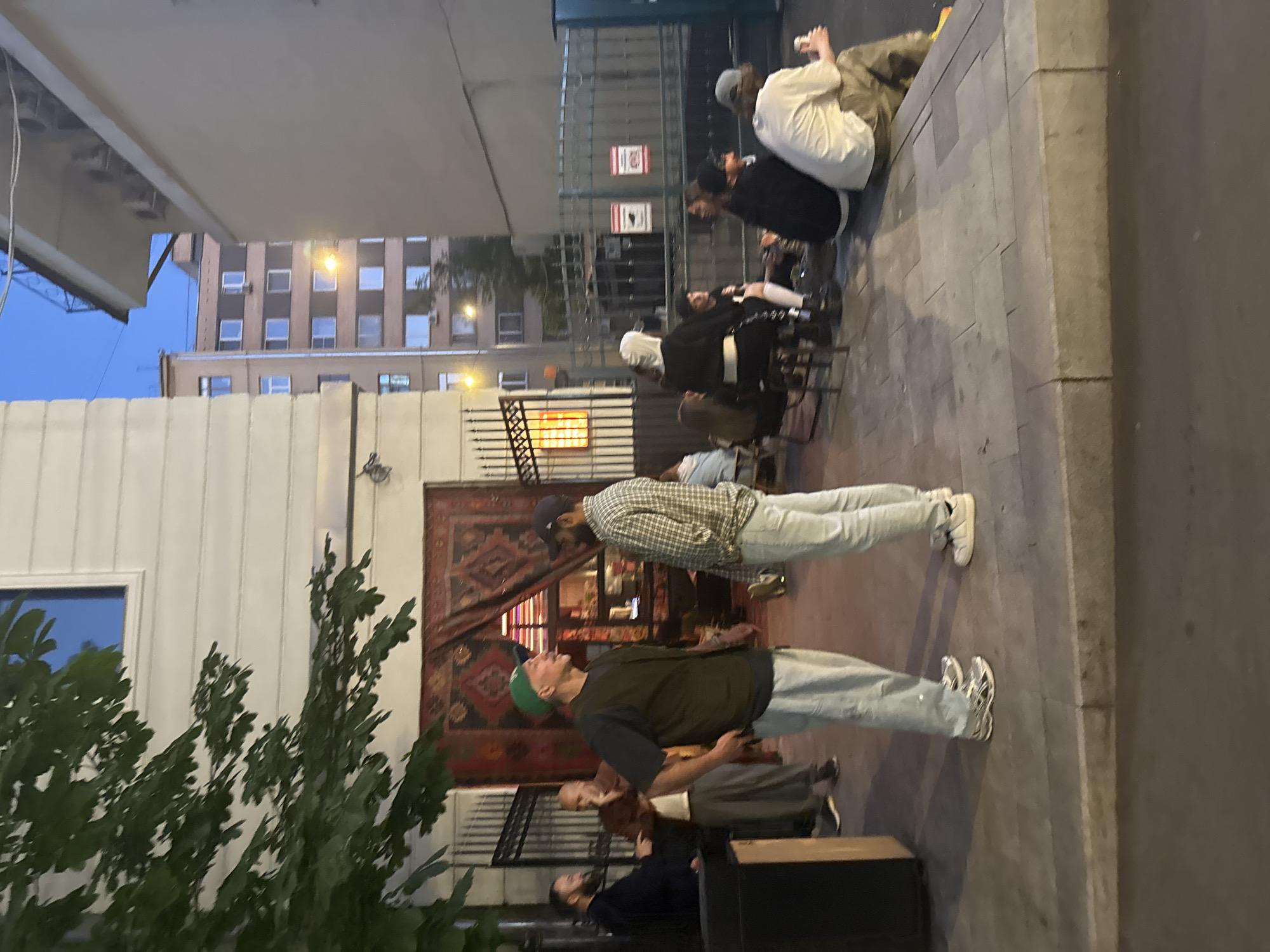 The image depicts an outdoor scene in Moscow, Russia, at dusk. In the foreground, two men are standing and conversing on a sidewalk. One man, wearing a green baseball cap, a dark vest, and light-colored pants, is facing to his right. The other man, in a plaid shirt and light-colored pants, is facing to his left. Behind them, a building with white siding and a large, patterned rug hanging on the exterior is visible. To the left of the building, a black trash receptacle is present. In the background, several other individuals are seated on chairs and the ground near a metal fence. The environment appears to be urban, with buildings and streetlights visible in the distance.