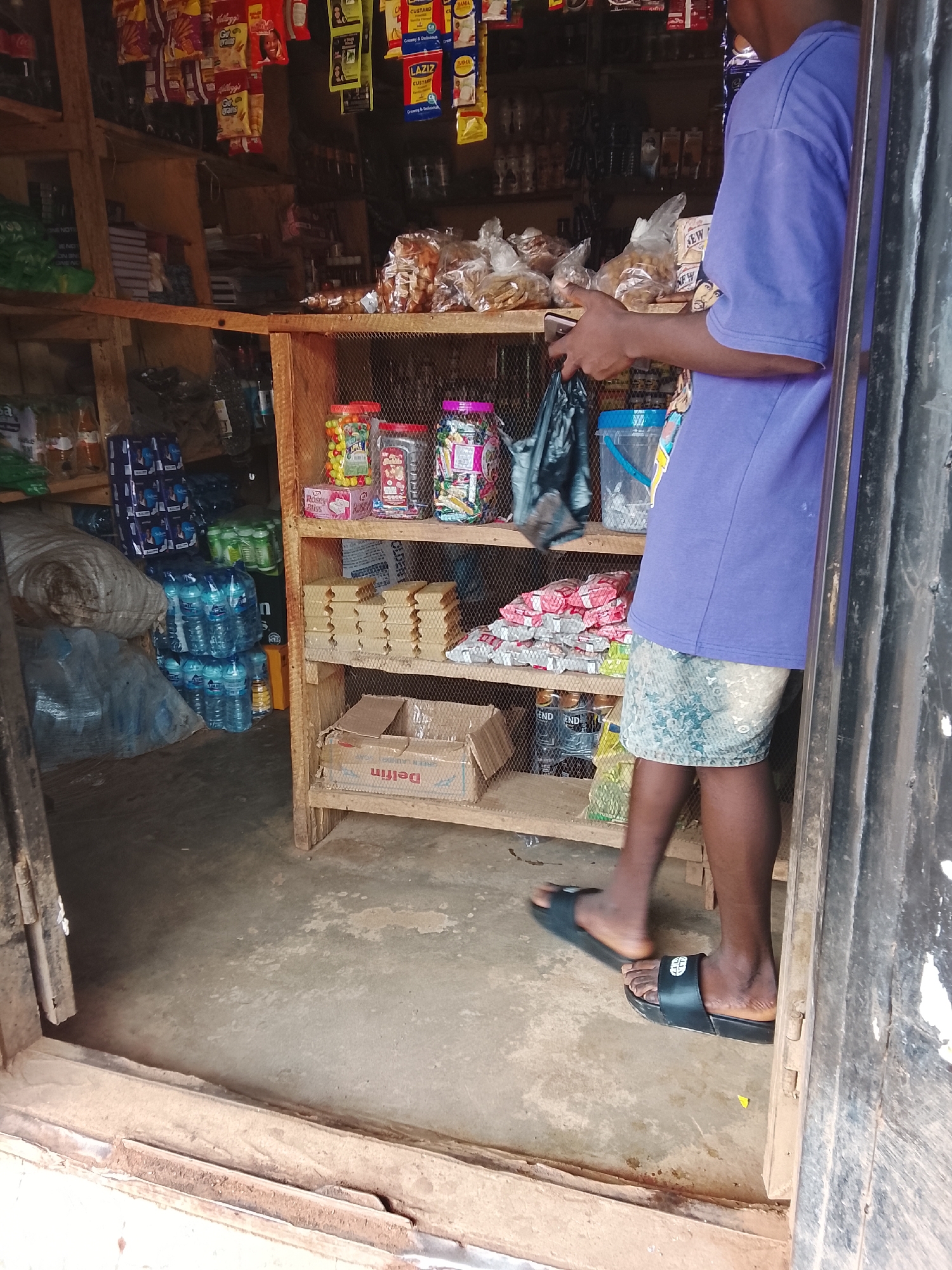 A person stands in a small, open-air shop in Durumi, Nigeria. The shop is stocked with various goods including bottled water, snacks, and other packaged items displayed on wooden shelves. The person, wearing a purple t-shirt and patterned shorts, is holding a mobile phone and a black plastic bag. The floor of the shop is made of concrete.