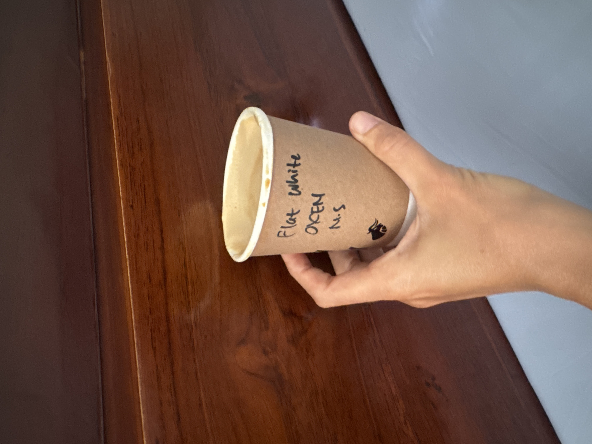 A close-up shot captures a hand holding a paper coffee cup against a dark, polished wooden background. The cup has handwritten text on it that reads "Flat White OXCE N N.S." and features a small black logo that appears to be an animal. The coffee inside the cup is light brown and slightly frothy. The wooden surface has a visible grain pattern and a rich, reddish-brown hue. The lower portion of the image shows a pale blue surface, possibly a table or counter. The lighting suggests an indoor setting, likely during the daytime.