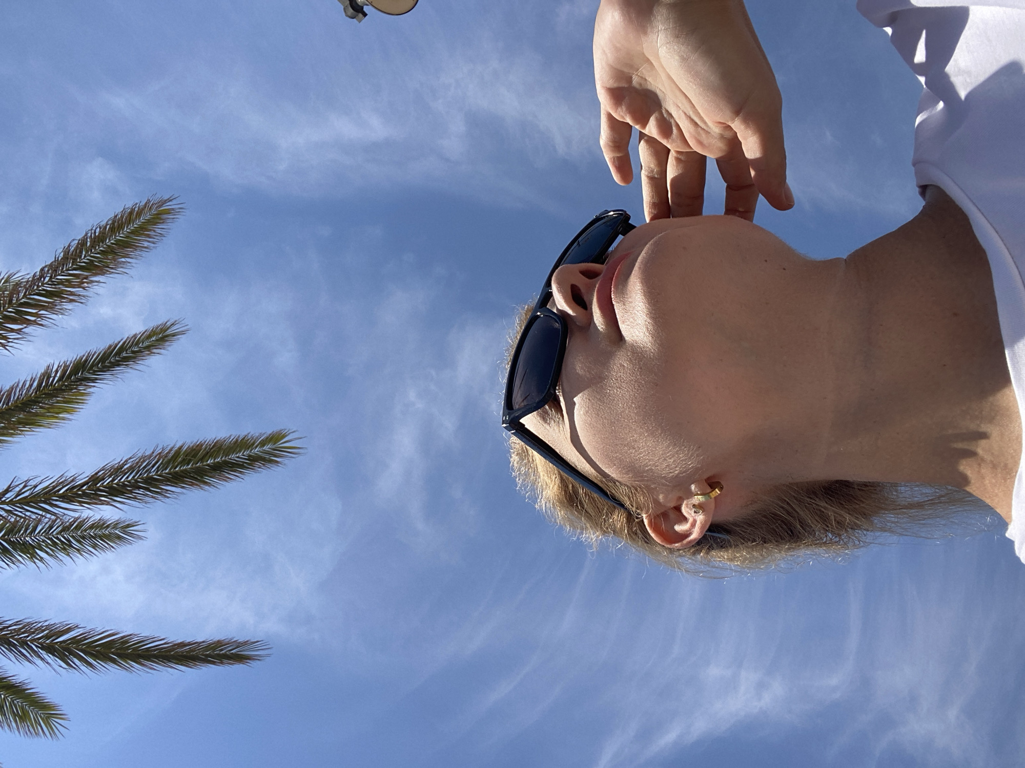 The image shows a close-up, low-angle shot of a person's head and shoulder against a bright blue sky with wispy white clouds. The person is wearing sunglasses and a white t-shirt. Their right hand is raised, with fingers lightly touching their forehead or temple area. A palm frond is visible in the lower left corner, suggesting a tropical or subtropical location. The sunlight is strong, casting shadows and highlighting the person's features. The time of day appears to be daytime, likely sunny. The person's expression is neutral or relaxed. The overall impression is one of leisure and outdoor activity in a warm climate. The location context is Valencia, Spain. No visible text is present.
