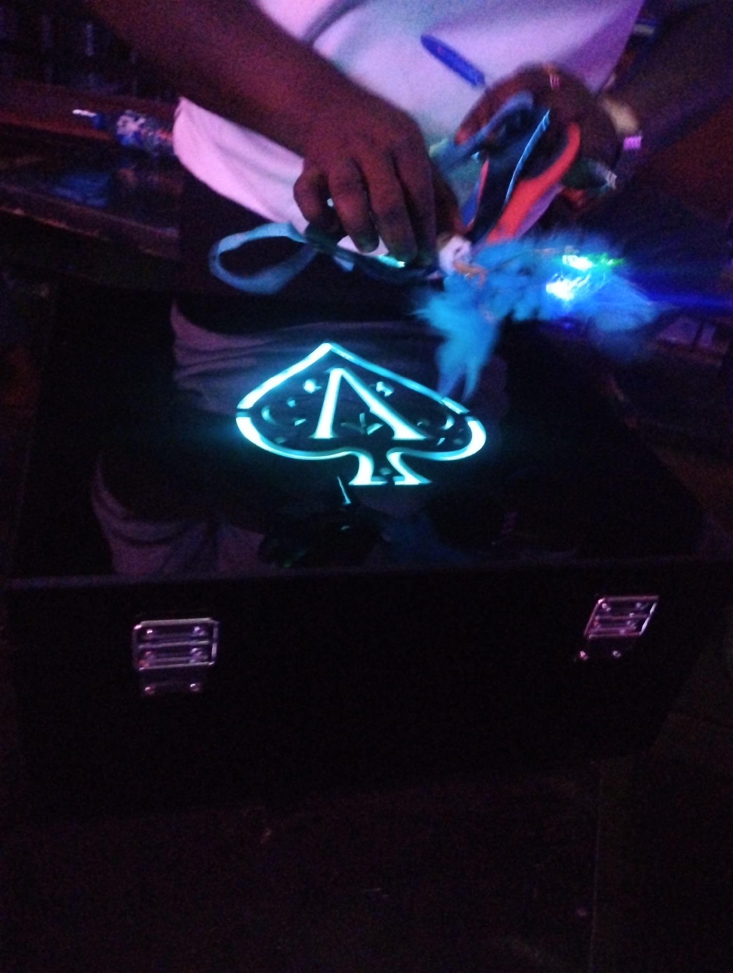 A person is seated in front of a dark, rectangular box with two metal latches on the front. The box is decorated with a neon blue spade symbol. The person's hands are visible, holding an item with blue feathers. The scene appears to be indoors, and the lighting is dim with a blue glow emanating from the spade symbol and the feathered object. There is not enough information to discern the city of Tumfure, Nigeria.