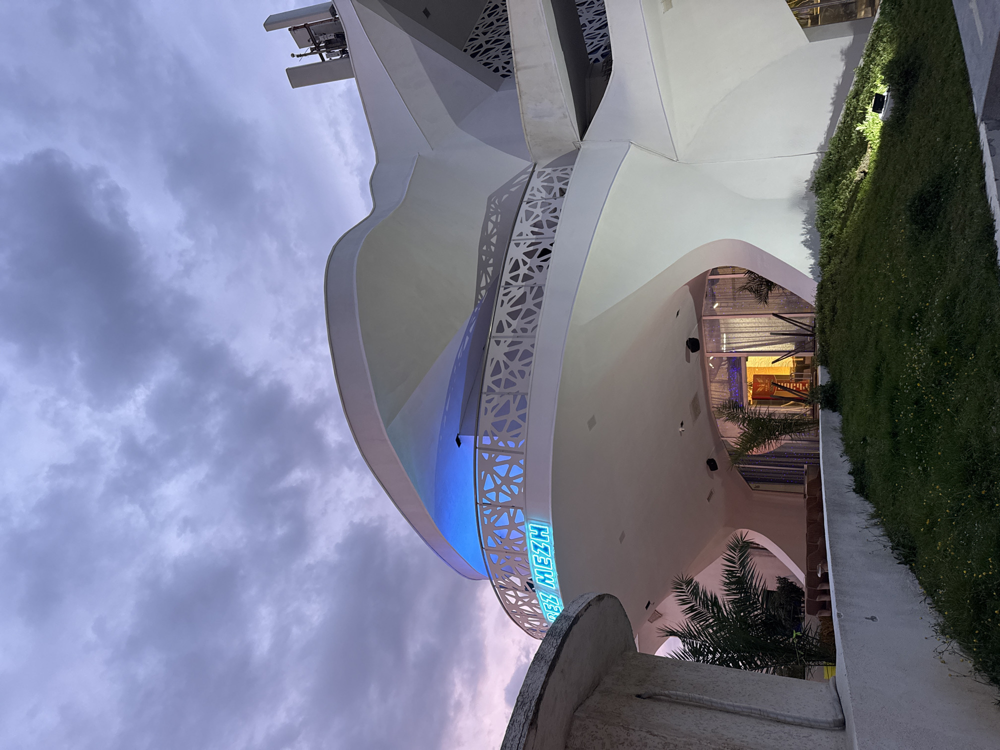 The image shows a building in Unknown, Unknown, from a low angle, tilted to the right. The building is primarily white, with a modern design incorporating curved shapes and geometric patterns. A blue neon sign, oriented vertically, is visible on the side of the building. The sky is cloudy with a mix of grey and pinkish hues, suggesting either sunrise or sunset. A green wall, resembling a vertical garden, is adjacent to the building on the right side. Palm trees are visible near the base of the building.