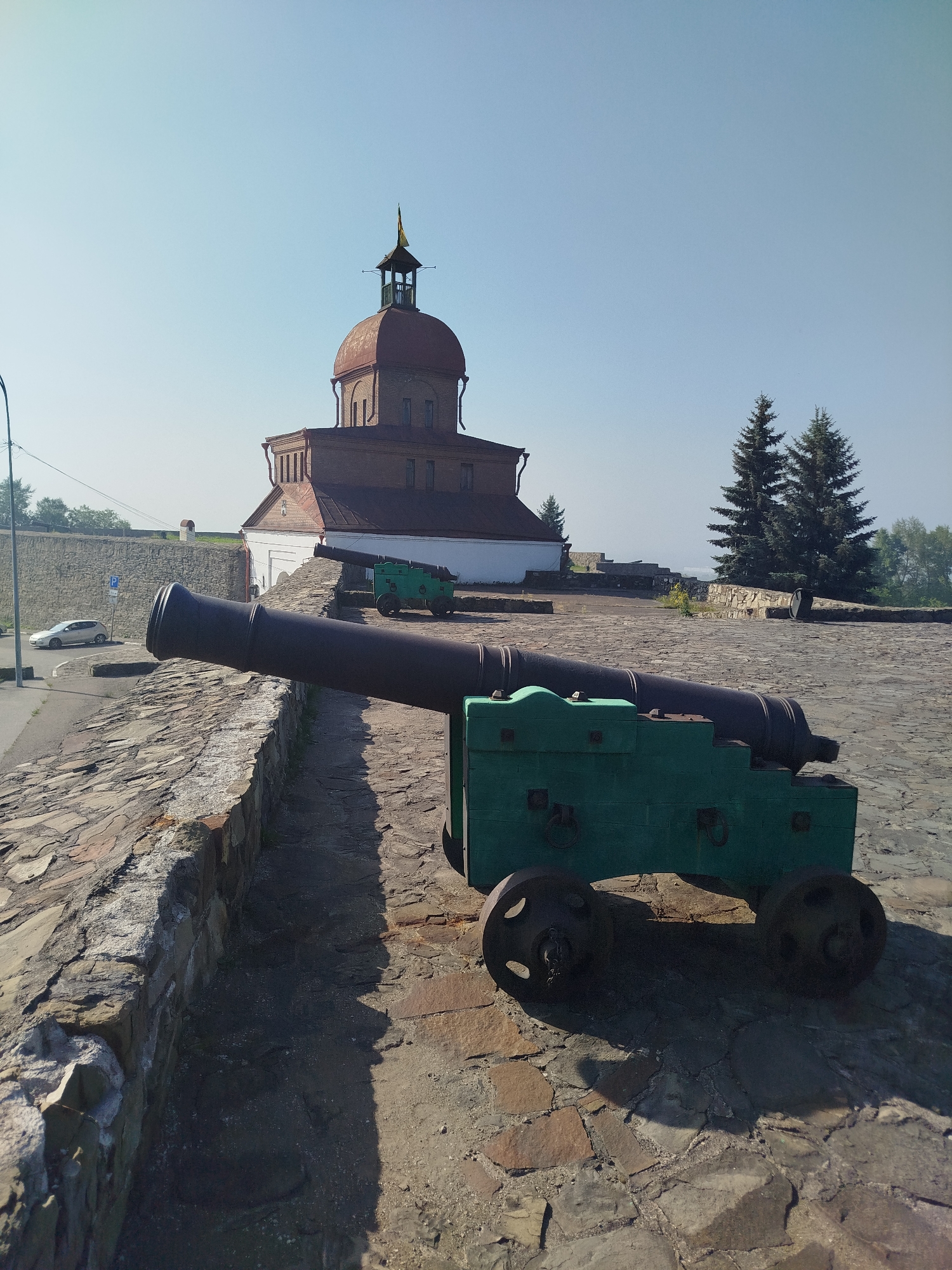 The image shows a stone fortress wall with two cannons on it. In the background, there is a brick building with a dome and a spire, and two tall evergreen trees. A white car is parked on a road next to the fortress wall. The scene is bathed in sunlight, casting shadows on the stone-paved ground. The location is the Novokuznetsk Fortress in Novokuznetsk, Russia.