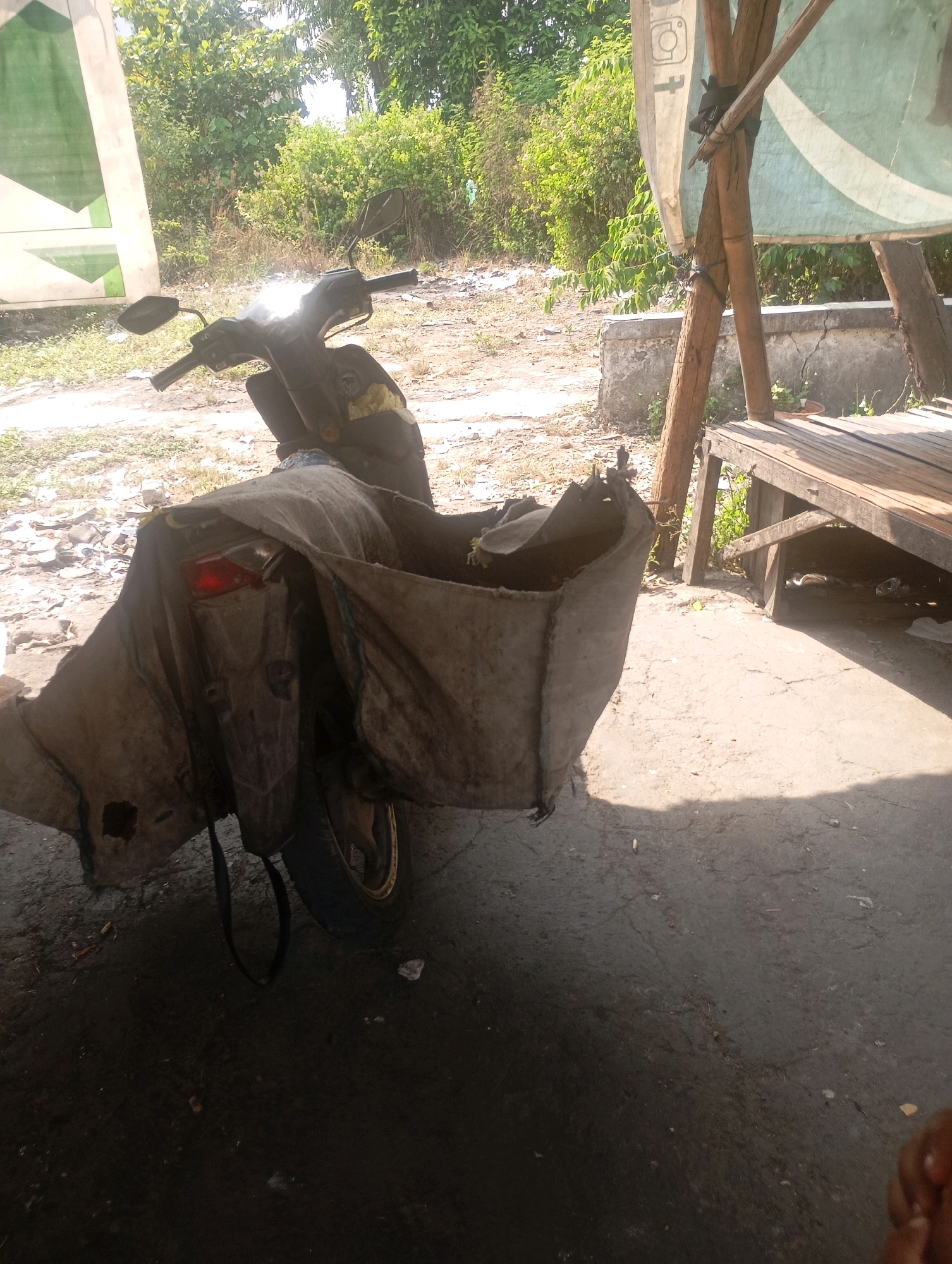 The image depicts a parked motorbike with a large, custom-made canvas bag attached to its rear. The bag appears to be made of a durable, weathered material and covers a significant portion of the back of the motorbike, extending downwards. The motorbike itself is dark-colored and has a single red taillight visible. It is parked on a paved surface, possibly a street or driveway, which is partially in shadow. In the background, there are trees and dense vegetation, suggesting an outdoor environment. To the right of the motorbike, a simple wooden bench is visible, supported by wooden posts and covered by a light-colored tarp. The scene has a rustic, outdoor feel, potentially in a residential or less developed area. The lighting suggests it is daytime, with sunlight casting strong shadows. The overall impression is of a utilitarian setup, perhaps for transporting goods. Based on the provided context, this scene is located in Babelan, Indonesia.