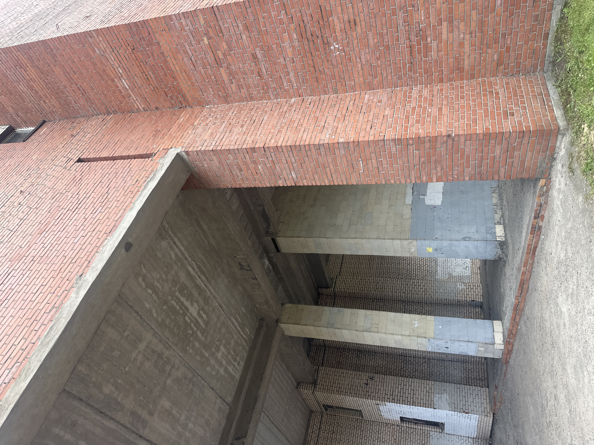 This media file depicts an outdoor scene of a modern architectural structure, likely under construction or undergoing renovation. The primary subjects are the building's exterior and interior elements, characterized by prominent use of red brick and concrete.
The exterior wall, constructed of uniformly laid red bricks, dominates the upper portion of the image. A section of the brickwork appears to be a cantilevered beam or walkway. Below this, the structure transitions to concrete, with large, unfinished concrete walls forming an open space. This open area reveals a multi-level interior with concrete pillars supporting what appear to be concrete beams or floors. The interior walls are also lined with brick, some lighter in color, creating a contrast with the red brick exterior.
There are no people or active events visible in the scene. The setting appears to be an urban or industrial environment, given the architectural style and materials. The presence of some sparse grass on the right side suggests an outdoor area adjacent to the building. The lighting indicates it is daytime, with diffused light suggesting an overcast sky or possibly early morning or late afternoon.
Notable details include the precise, almost repetitive patterns of the brickwork, and the raw, unfinished nature of the concrete. There is some minor debris or discoloration on some of the concrete surfaces and a few small, dark lines embedded in the brickwork, possibly structural elements or markings. A small, dark rectangular window opening is visible in the background brickwork. No discernible text is present in the image. The overall impression is one of an unpopulated, monumental structure.