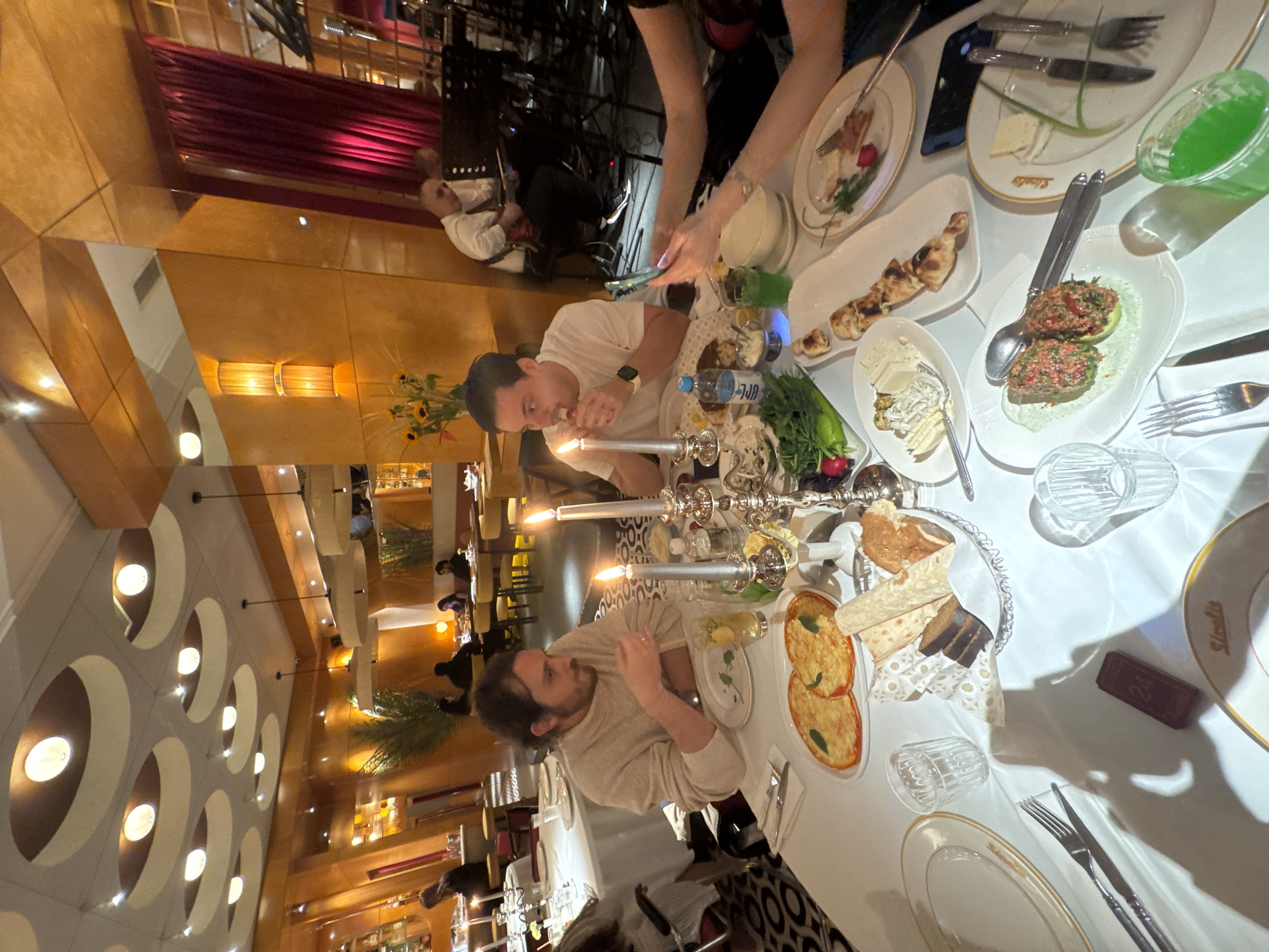 The image captures a dining scene at a restaurant in Yerevan, Armenia. A man is seated at a table covered with a white tablecloth, laden with plates of various foods, likely part of a shared meal. A woman stands nearby, using her phone to take a photo. Silver candelabras with lit candles add to the ambience. In the background, a musician plays an instrument, possibly a violin. The interior has a warm lighting scheme, with a patterned ceiling and wall decor. The scene depicts a social gathering, most likely a dinner or celebratory event, in an upmarket or traditional Armenian restaurant setting.