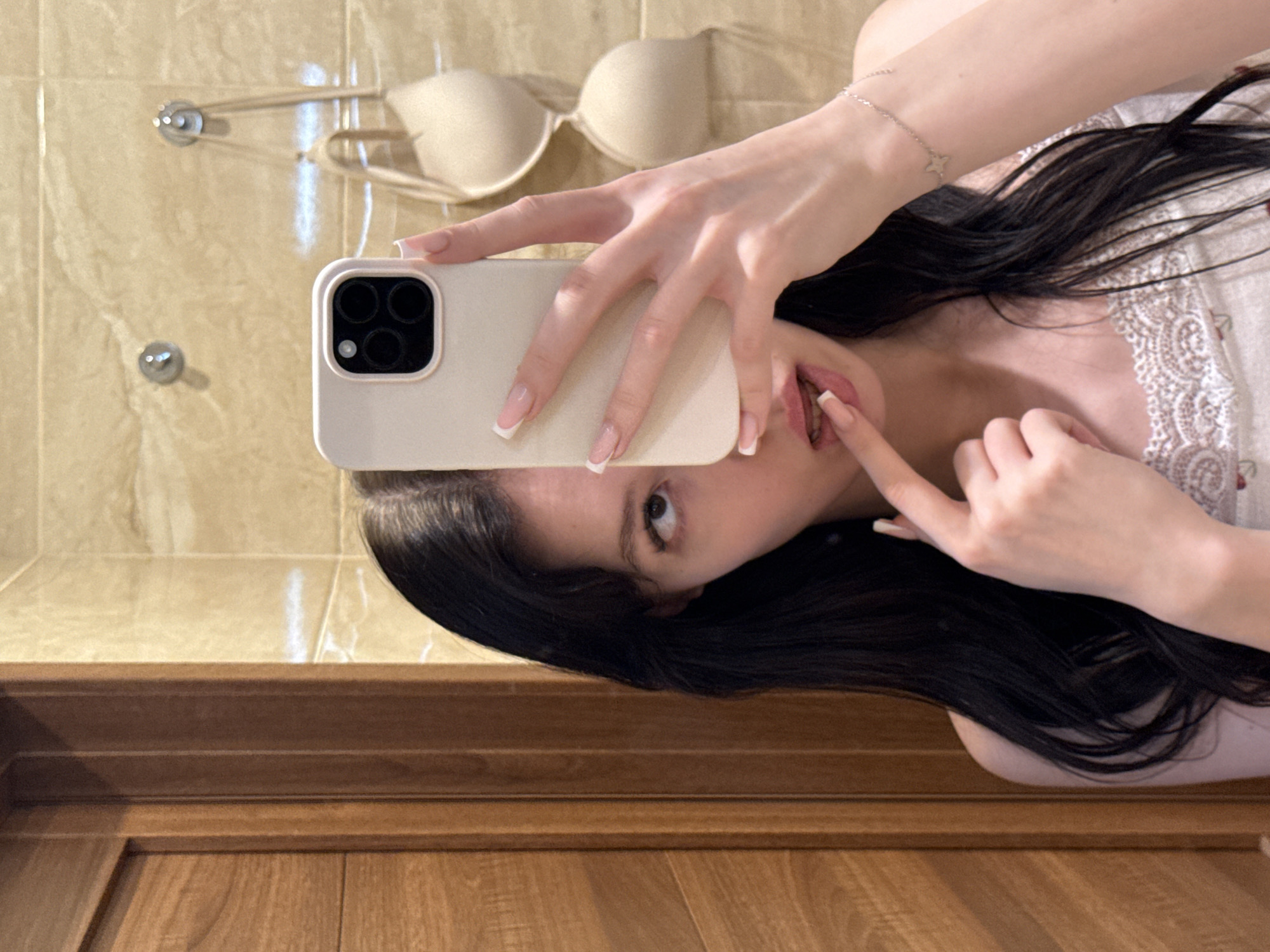 A young woman with long, dark hair is taking a selfie in a bathroom. She is holding a light-colored smartphone in her right hand, obscuring part of her face. Her left index finger is touching her lips. She is wearing a white top with lace trim and a cherry pattern. In the background, a beige bra is hanging on a hook. The walls are tiled with a beige, marble-like pattern, and there is a wooden door to the left of the frame. The image appears to have been taken in a residential bathroom in Khimki, Russia.