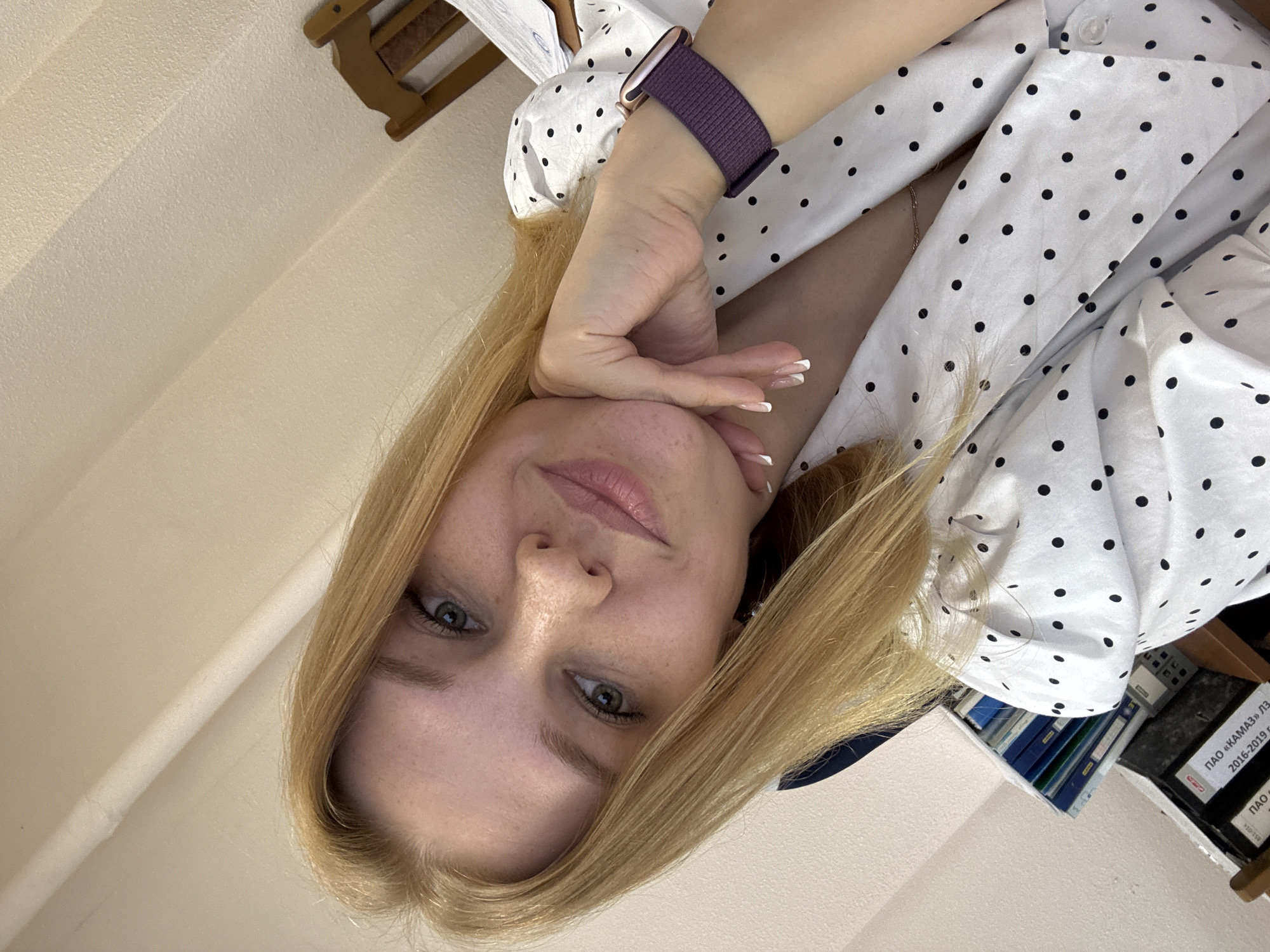 A selfie photograph shows a woman with blonde hair and light skin, resting her head on her hand, which wears a purple smartwatch. She is wearing a white shirt with black polka dots. The background includes a light-colored wall, a portion of a wooden chair, and a bookshelf with blue binders and files. A document with Russian text is visible. The location, Rusanovo, Russia, is not directly identifiable from the image alone.