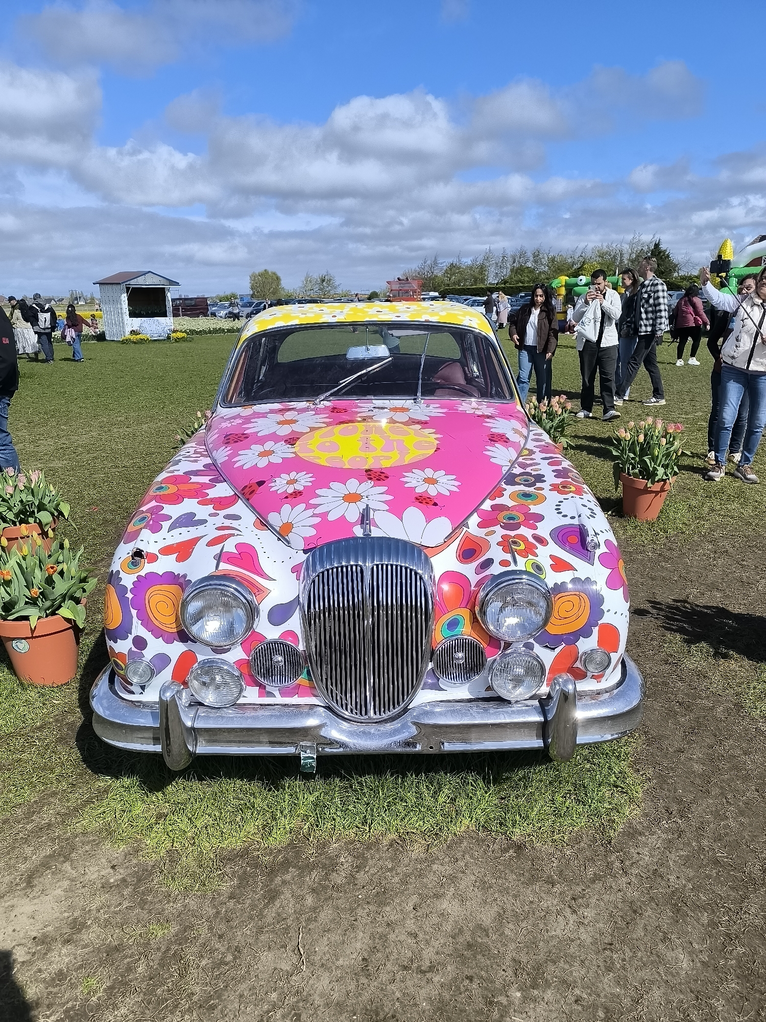 The image depicts a flower-painted vintage car parked on a grassy field, likely a tulip field based on the presence of tulip bulbs in pots. The car features a white base color with psychedelic patterns including flowers, hearts, and swirls, predominantly in shades of pink, yellow, purple, and orange. It has a chrome grill and bumper, with a yellow-painted roof. People are gathered in the background, some taking pictures. A small white building is visible in the left background. The sky is blue with scattered white clouds, suggesting a partly sunny day. The location appears to be a tulip farm in Hillegom, Netherlands, based on the common association of tulips and flower fields with the region.