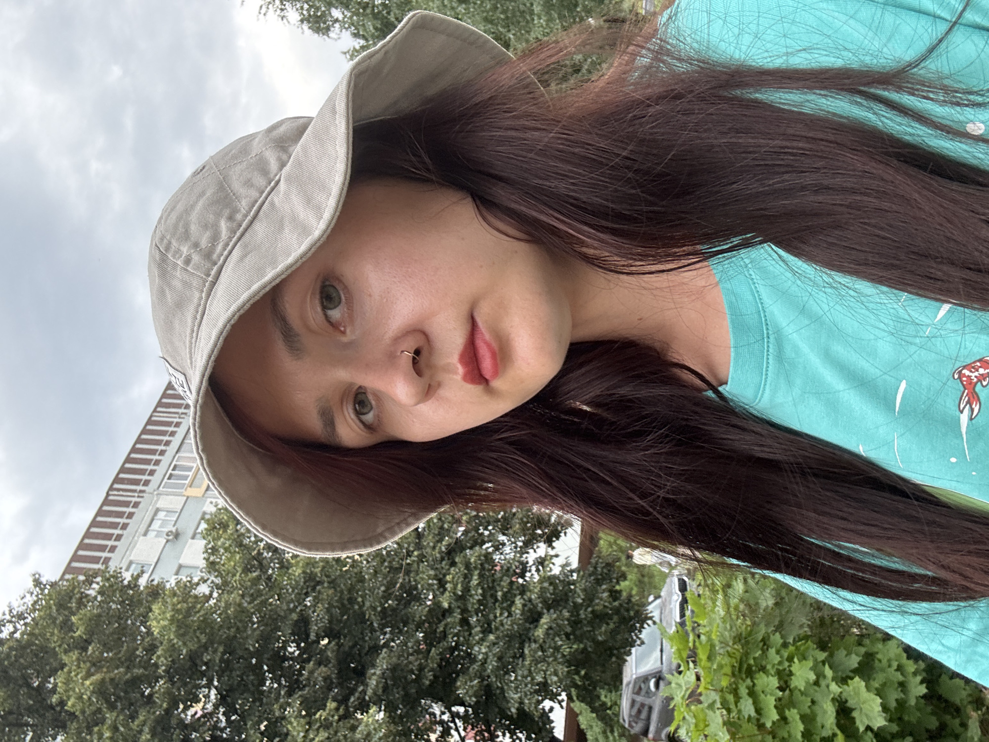 A medium shot portrait of a young woman wearing a light gray bucket hat and a turquoise t-shirt with a graphic print. She has dark brown hair, a nose ring, and is looking directly at the camera. The background is an outdoor urban setting with a tall apartment building visible in the distance, along with trees and a car parked on the street. The sky is overcast. This scene is located in Naberezhnye Chelny, Russia.