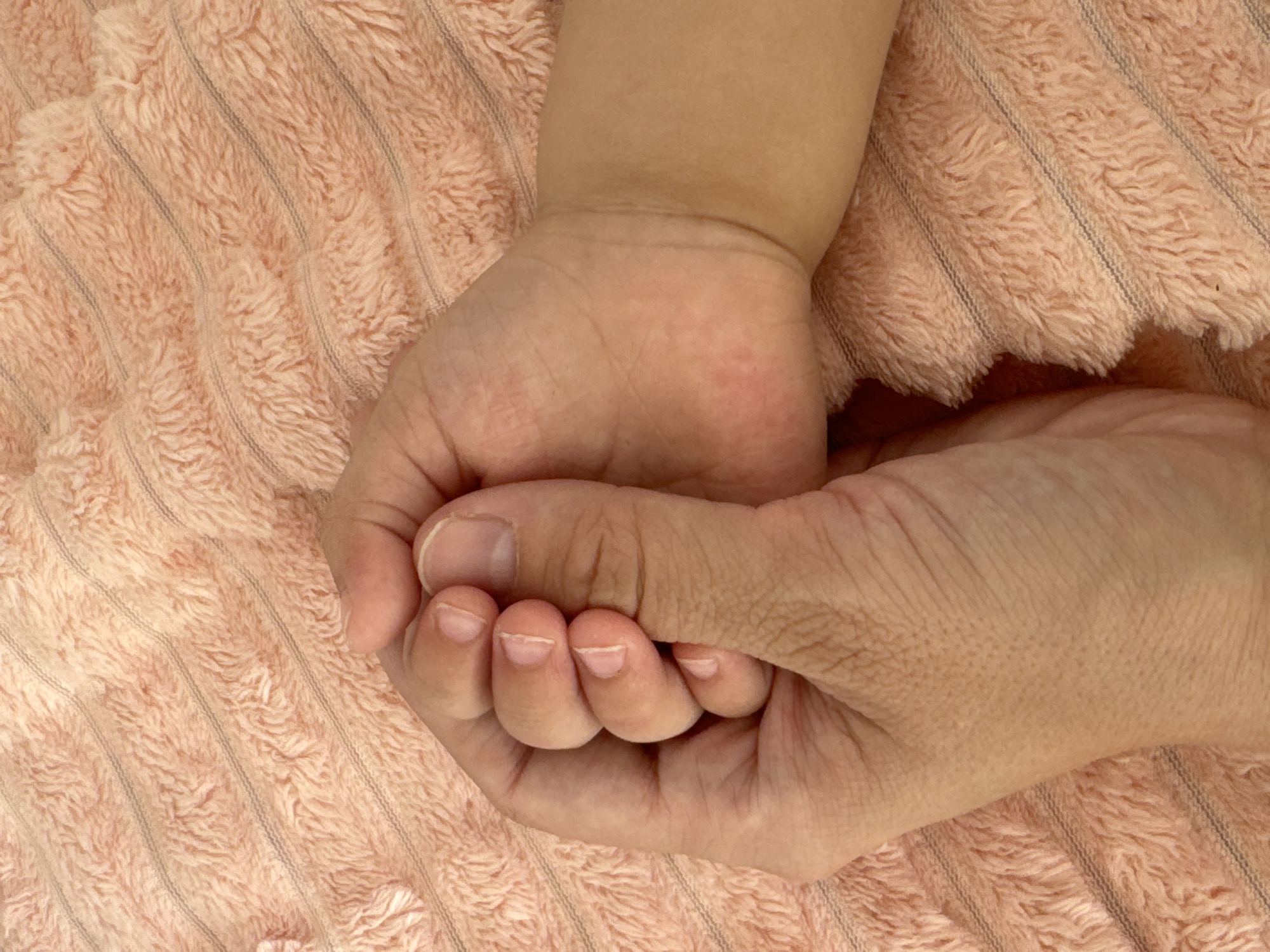 A close-up shot depicts an adult hand holding a baby's hand against a peach-colored, striped fabric background. The adult's hand is slightly wrinkled, while the baby's hand appears smooth. No other elements are discernible from the image. No information about the location in the city of Chaikovsky, Russia, is provided within the image.