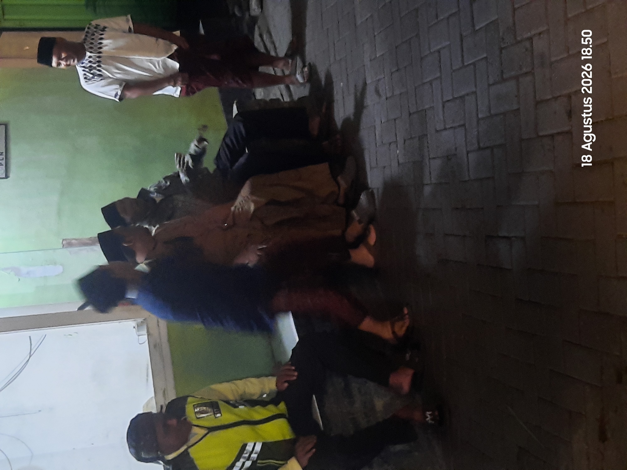 The image depicts an outdoor scene with four individuals gathered on a paved walkway. The setting appears to be in Sukokerto, Indonesia.
In the foreground, on the left, a person wearing a bright yellow and green vest over black pants is seated. Their face is obscured by shadow.
To the right of the seated individual, three other people are visible. Two of them are seated, and the third is standing. The two seated individuals are wearing traditional Indonesian attire, including peci caps. The standing individual, on the far right, is wearing a white short-sleeved shirt with a geometric pattern across the chest and dark red pants. This person is looking to their left and smiling.
The environment includes a light-colored wall in the background, with some visible wires and a framed sign overhead. The ground is paved with interlocking bricks. The lighting suggests it is either dusk or nighttime, with artificial light sources illuminating the scene. The overall atmosphere is casual and social.
A timestamp is visible in the lower right corner of the image: "18 Agustus 2026 18.50".