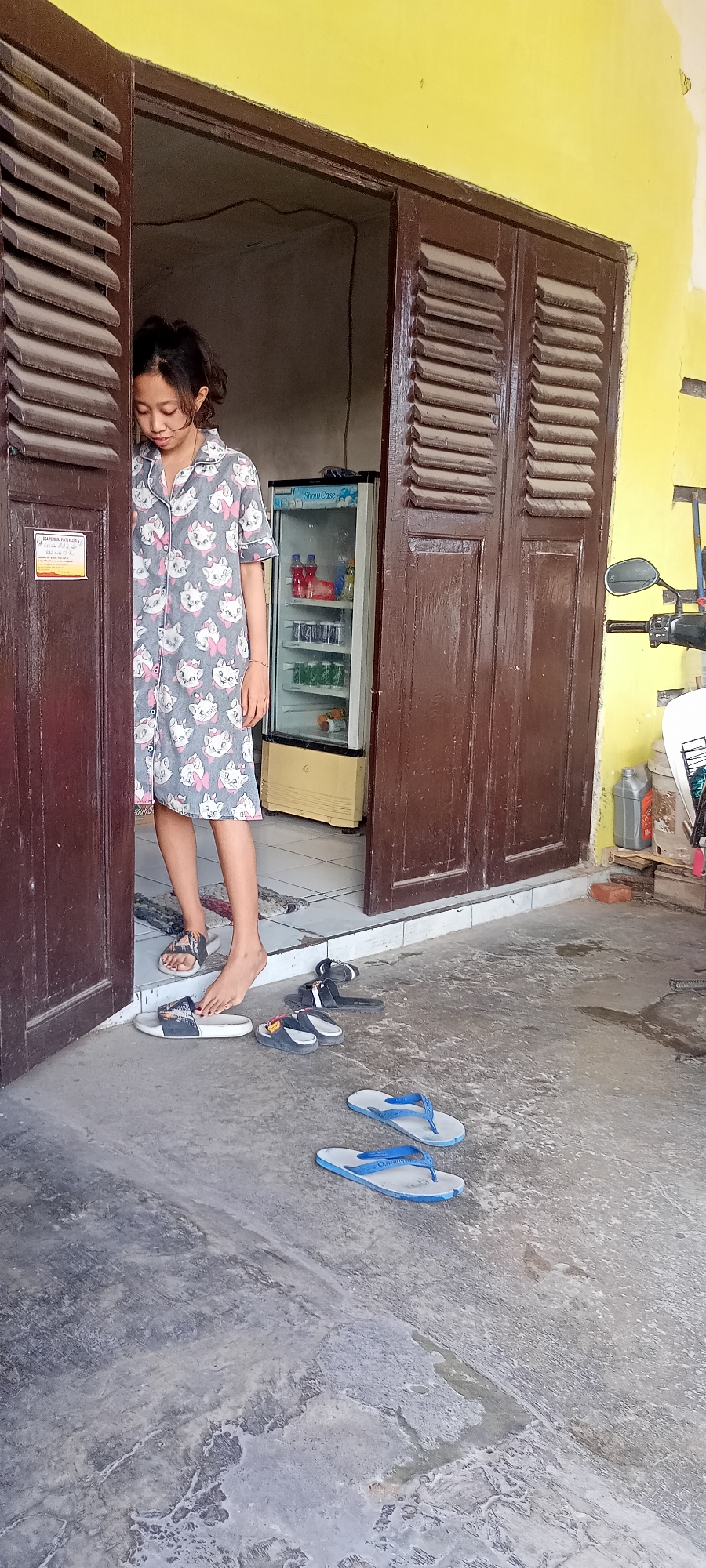 A young girl with dark hair pulled back in a ponytail stands in a doorway, looking down. She is wearing a grey nightgown with a repeating pattern of white cartoon cats and pink bows. The girl is standing on a tiled floor with a rug visible behind her. In front of her, on the concrete ground, are several pairs of footwear, including white slides with a black stripe, dark flip-flops, and blue flip-flops. To the right of the doorway is a tall, narrow glass-fronted refrigerator with a yellow base, containing bottles and cans. The doorway is framed by dark brown wooden shutters. The building appears to be a shop or residence located in Gempolklutuk, Indonesia, with a yellow-painted wall visible to the right. A portion of a motorcycle is visible on the far right.