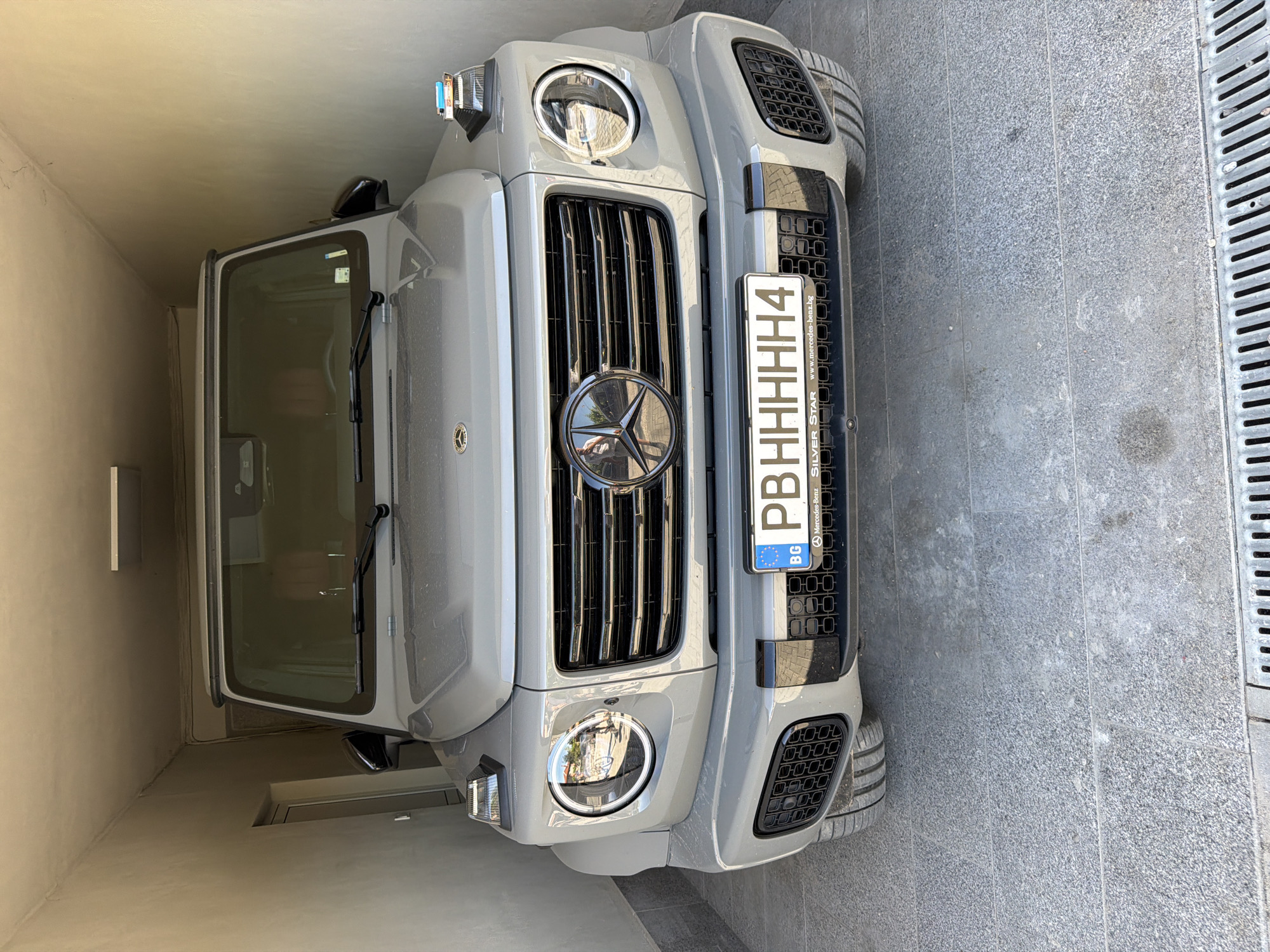 The image shows the front of a gray Mercedes-Benz G-Class parked in what appears to be a tight alleyway or garage in Asenovgrad, Bulgaria. The vehicle is facing the camera. The front grille, adorned with the Mercedes-Benz logo, is prominently visible. The license plate, with the Bulgarian country code "BG," reads "PBHHHHH4." The text "SILVER STAR" and "www.mercedes-benz.bg" are on the license plate frame. The vehicle is parked on a gray stone surface near a narrow drain cover with parallel openings. The alleyway walls are beige.
