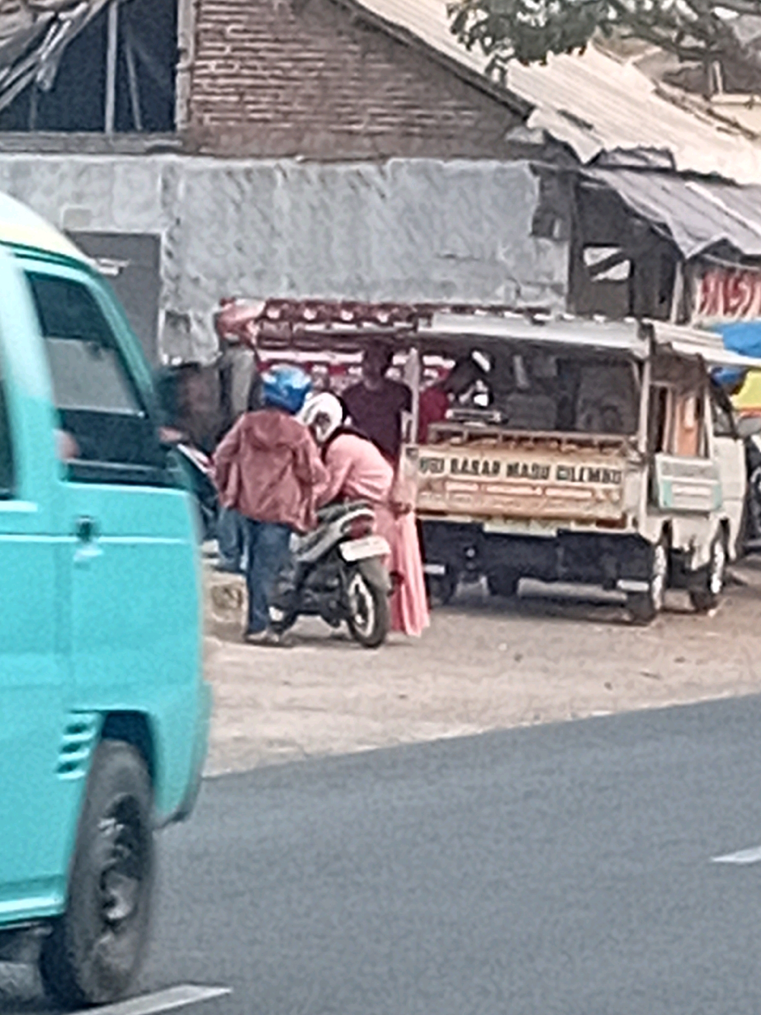 The image depicts a street scene in Besito, Indonesia. In the foreground, a light blue van is partially visible on the left. In the middle ground, two individuals are interacting near a motorcycle parked on the right side of the road. One person, wearing a blue helmet and a reddish-brown jacket, is standing next to the motorcycle. Another person, wearing a pink garment, is seated on the motorcycle. Behind them, a small truck with a wooden bed is parked. The truck has writing on its side, partially obscured. In the background, there are buildings with corrugated metal roofs and brick walls, suggesting an urban or semi-urban environment. The ground appears to be asphalt.