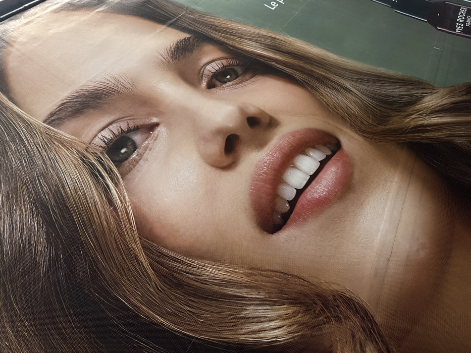 The image displays a large-format advertisement featuring a close-up of a young adult female's face, observed in the city of Paris, France. The woman has light brown, wavy hair framing her face, fair skin, and green eyes that gaze slightly upward and to the right. Her eyebrows are well-defined, and her lips are full and subtly colored pink, parted in a slight smile revealing white teeth. The background behind her is a solid dark green. In the lower right corner, branding for "YVES ROCHER FRANCE" is visible in white text, partially overlaying a dark bottle with a dropper applicator. The overall image exhibits the texture of a printed banner or billboard material, with minor creases visible across the surface.
