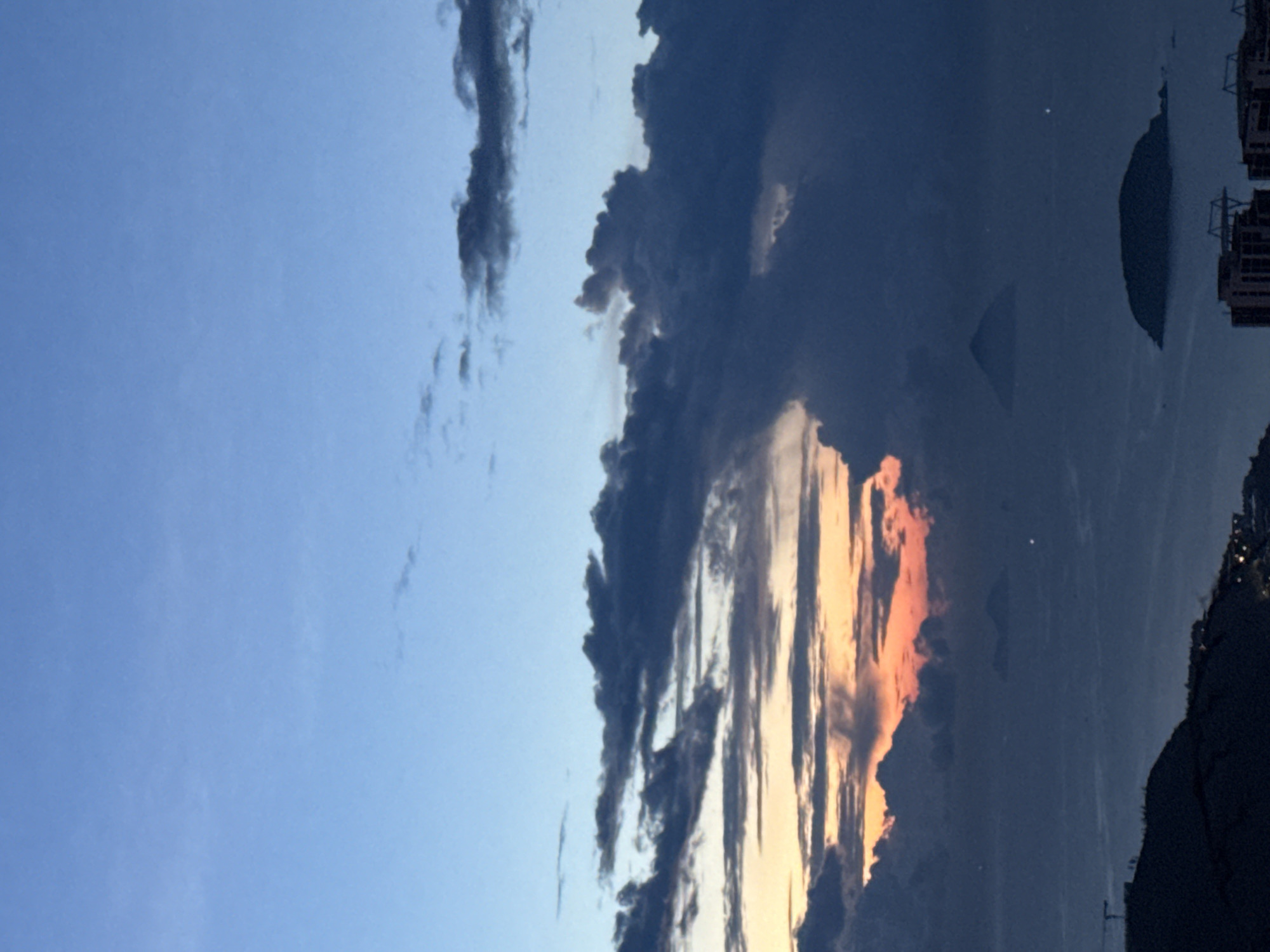 The image shows a vertical landscape of a dusk scene. The sky transitions from a pale blue on the left to darker, cloudier regions toward the right. Prominent dark clouds dominate the right side of the frame, some with orange and yellow hues along their lower edges, indicating the setting sun. In the distance, a body of water reflects the sky, with landmasses and city lights faintly visible. A dark landmass, possibly a mountain or hill, is visible on the right side of the scene, and the overall impression suggests a coastal location, possibly in Bac Nha Trang Ward, Vietnam.