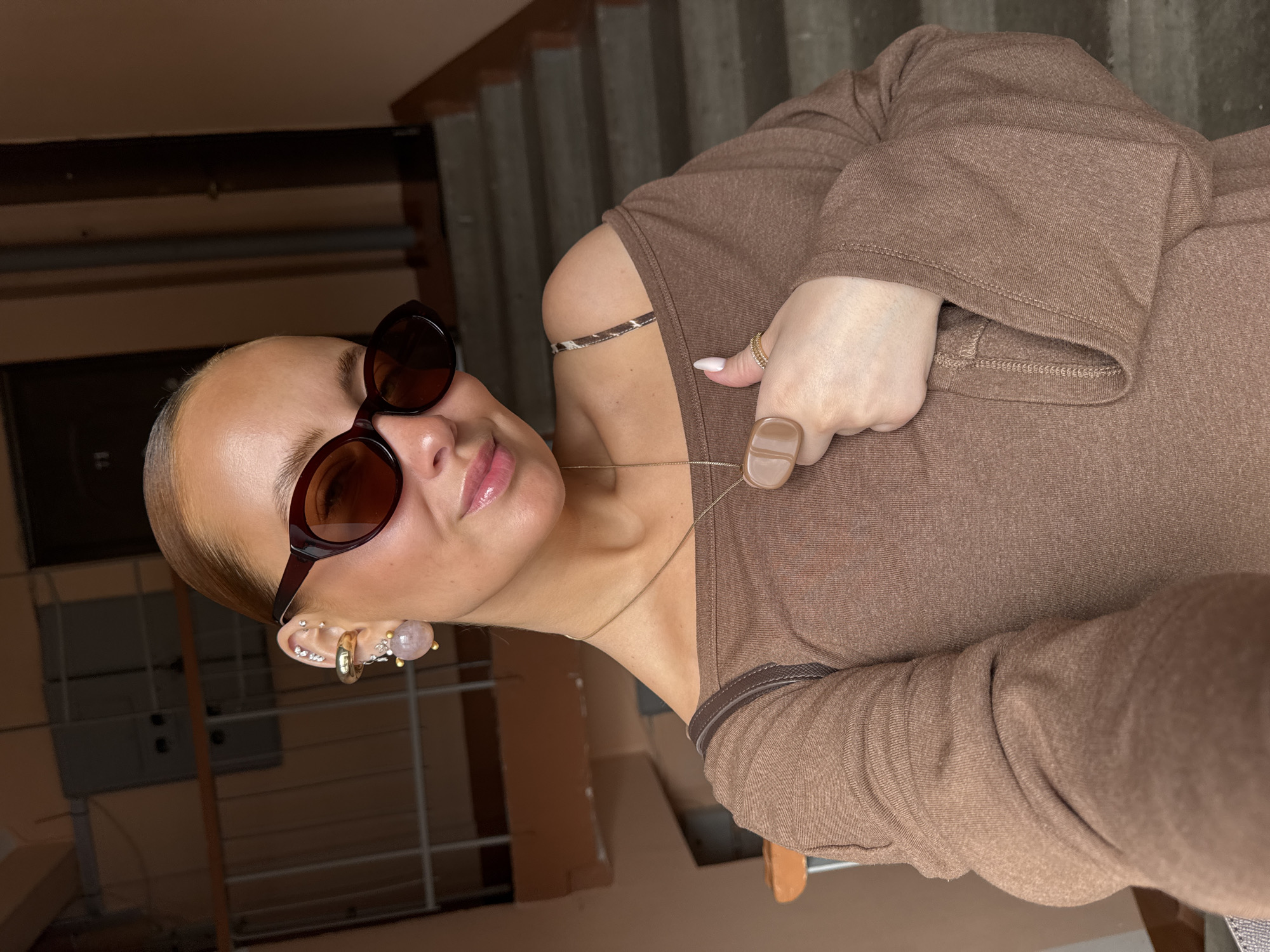 A woman stands indoors, likely in a stairwell based on the background of concrete steps and railings. She is wearing brown-tinted sunglasses and multiple earrings in her left ear, including hoops, studs, and a larger dangling earring with a pink sphere. The woman is dressed in a brown, long-sleeved top with a design that exposes one shoulder. She is wearing a thin necklace. Her nails are manicured with light-colored polish. On her right hand, she wears a gold ring and a large, rectangular ring. The location is likely in Chaikovsky, Russia.
