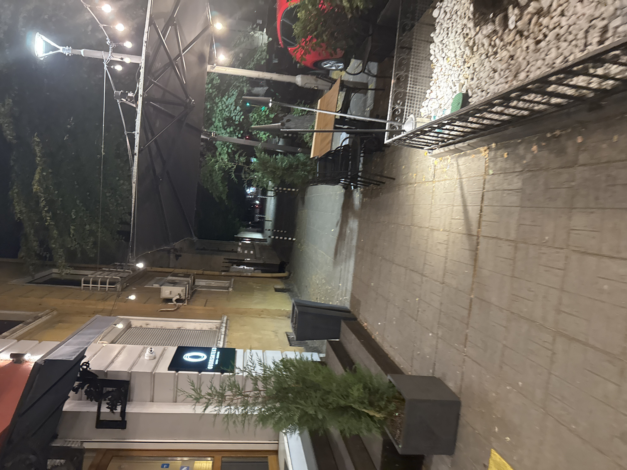The image depicts the exterior of a building, likely a restaurant or cafe, at night. A sidewalk runs along the front of the building, with a paved walkway. Several potted plants are visible, including a tall evergreen tree in a square planter near the entrance. The building facade is a pale yellow color, with an awning over the entrance. A sign with a circular logo and text is mounted on the wall. Outdoor seating is arranged on the sidewalk, with tables and chairs under a large umbrella. String lights and a lamppost illuminate the area. A red car is parked nearby, and trees and foliage are visible in the background. The scene suggests a European urban setting, likely Sofia, Bulgaria, given the architectural style and street furniture.