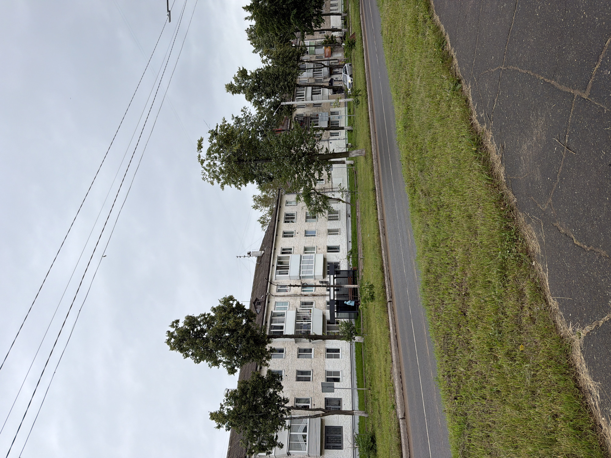 The image depicts a street scene in the city of Chaikovsky, Russia, on an overcast day. A row of multi-story apartment buildings, constructed with light-colored textured material, lines the street. Trees with full green canopies are planted in front of the buildings. A paved road runs horizontally across the foreground, with a grassy verge separating it from the sidewalk and buildings. There are power lines stretching across the sky. A bus stop shelter is visible in front of one of the buildings, with a single person in a blue jacket standing inside. A silver car is parked on the side of the road. The overall impression is of a typical urban residential area.